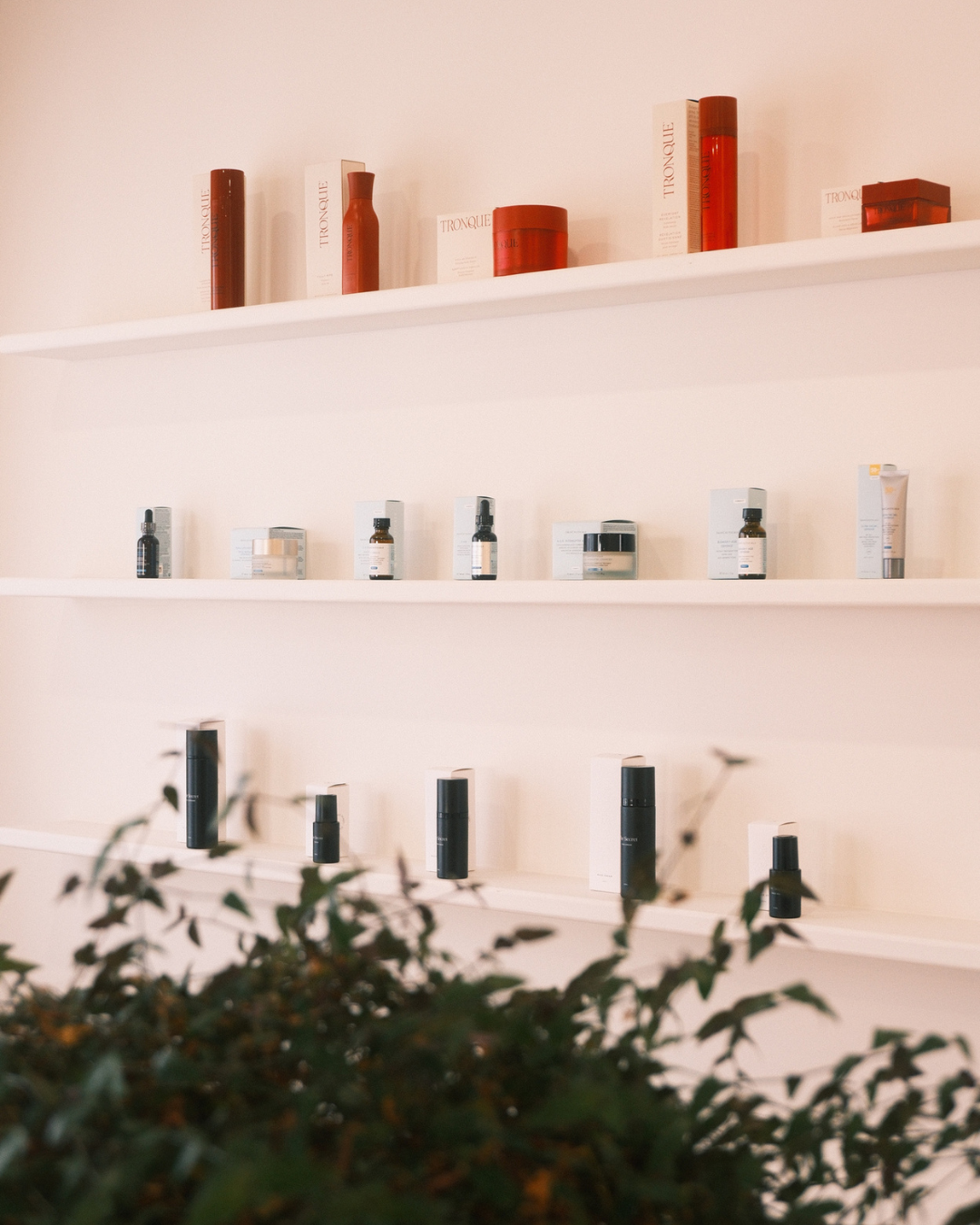 Clean display of skincare and beauty products on white shelves against a light pink wall, with blurred greenery in the foreground.