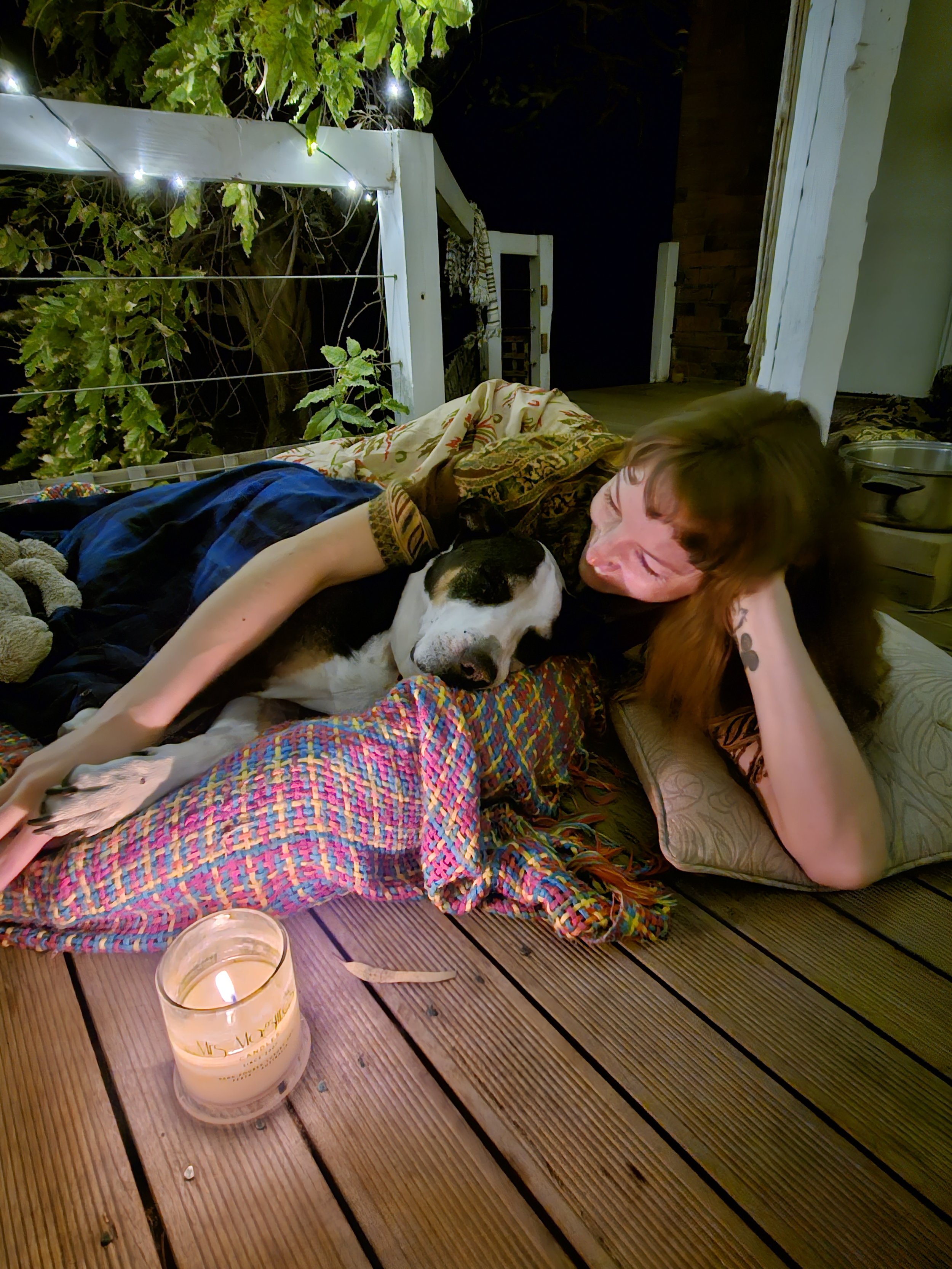 A person lying on a wooden outdoor deck at night, with a dog resting on their chest, near a lit candle and a glass. There are trees with green leaves, string lights, and a white fence in the background.