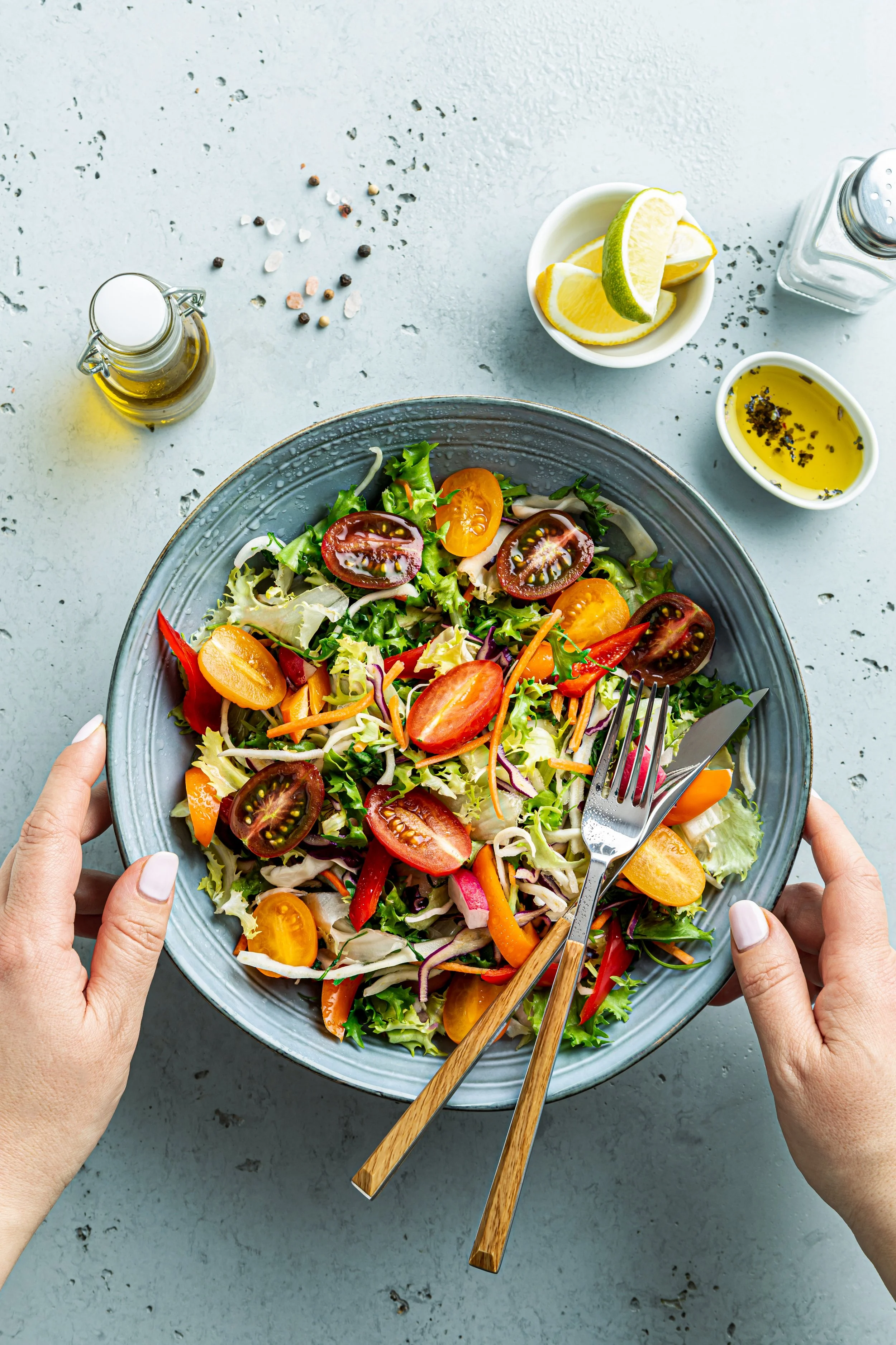 A colorful mixed salad with cherry tomatoes, red bell peppers, carrots, and lettuce, held in a gray ceramic bowl with a fork and spoon. Hands are visible holding the bowl, on a light gray textured surface with lemon slices, olive oil, pepper, and salt shakers nearby.