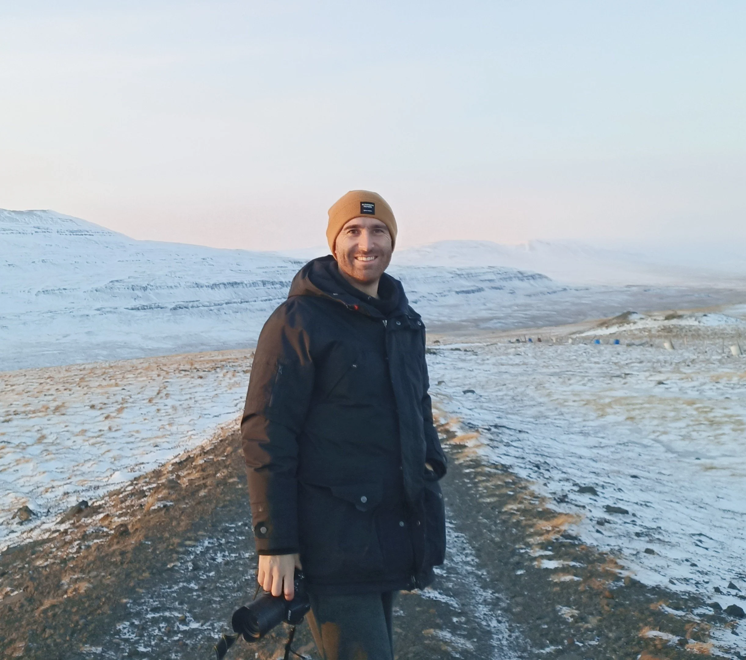 Homme souriant portant un chapeau brun, une veste noire et tenant des jumelles, debout sur un chemin enneigé dans un paysage de montagnes enneigées au loin, lors d'une journée ensoleillée.