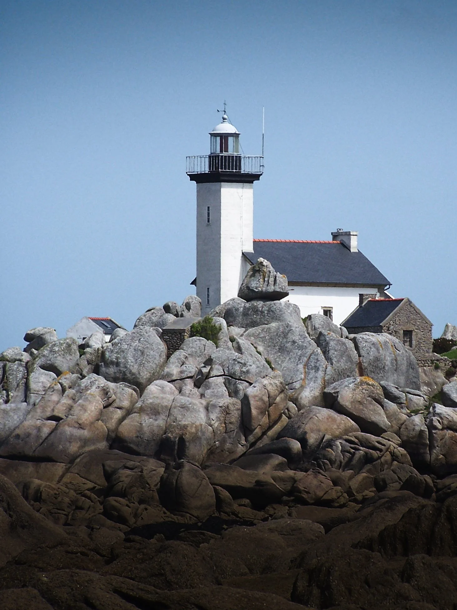Phare blanc situé sur des rochers, avec un bâtiment adjacent, contre un ciel clair.