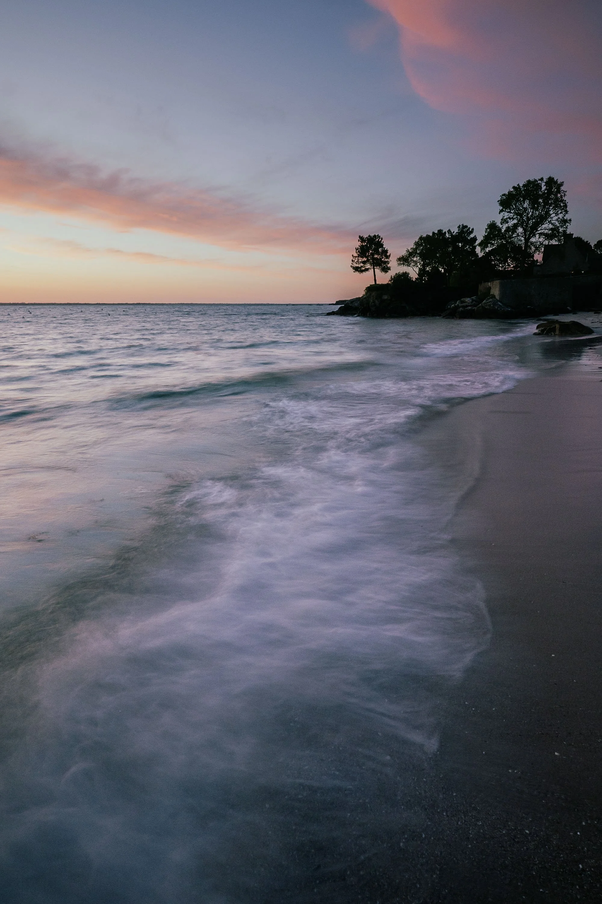 Plage au crépuscule avec vagues douces, rochers, arbres et maison en silhouette sous un ciel rose et violet.