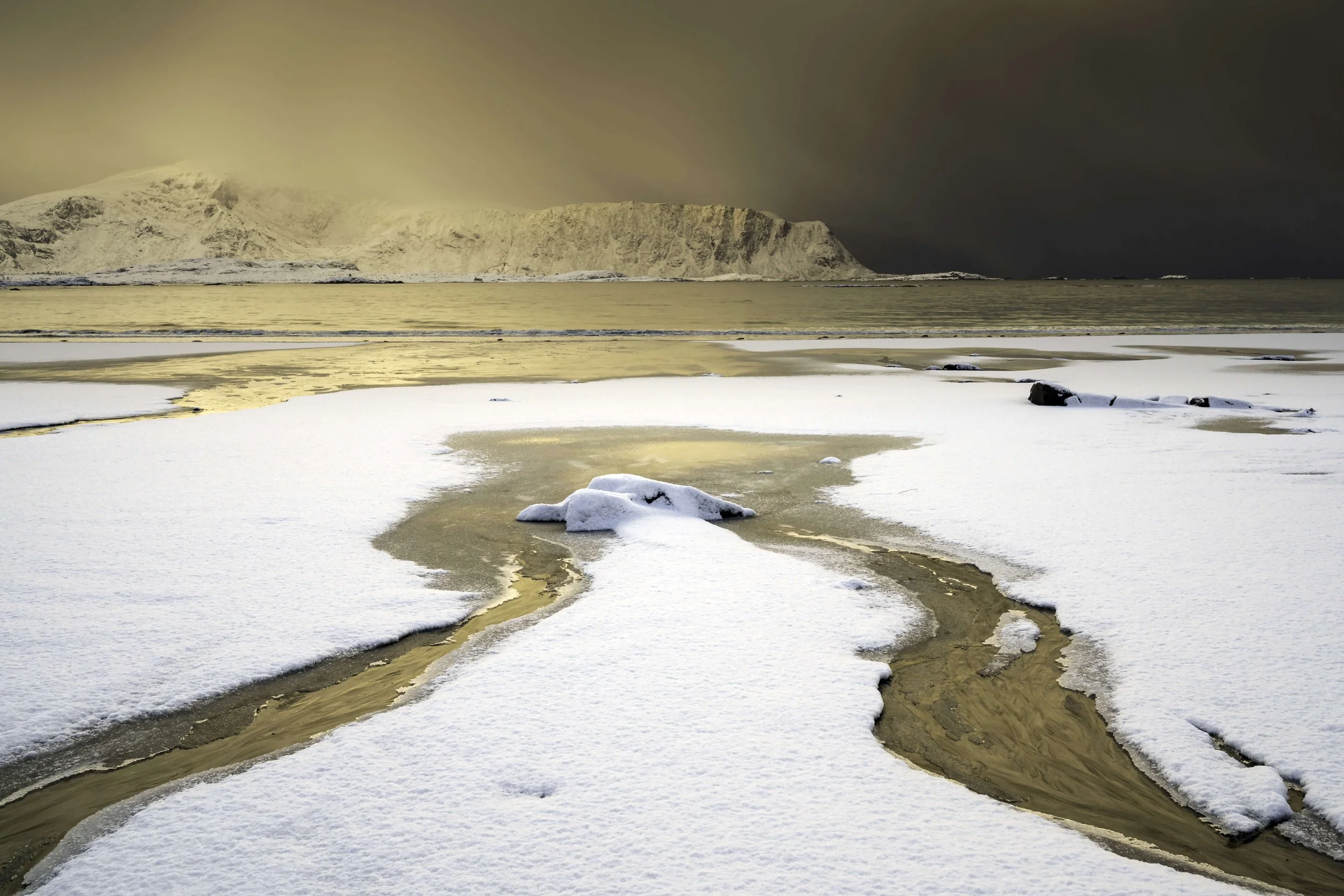 Paysage de neige avec un rivage gelé, une mer calme et une montagne enneigée en arrière-plan sous un ciel sombre.