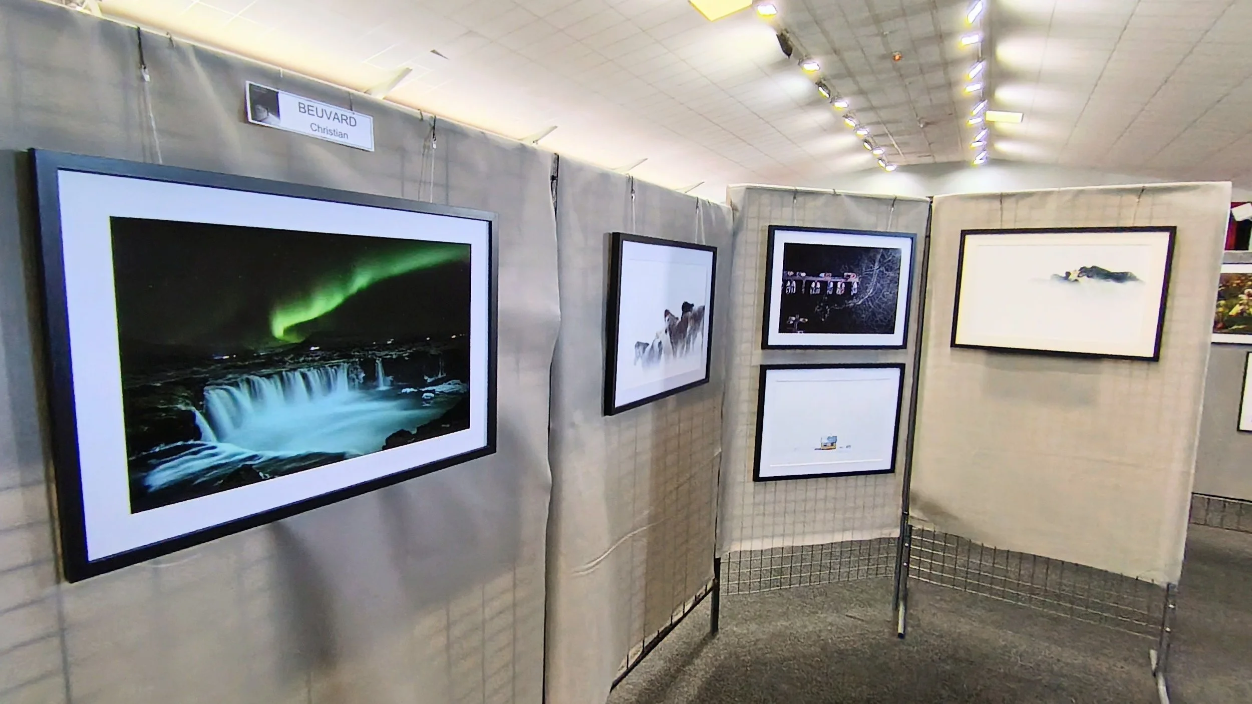 Exposition de photos avec plusieurs cadres suspendus sur des cloisons en toile, représentant des paysages naturels et des phénomènes comme l'aurore boréale.