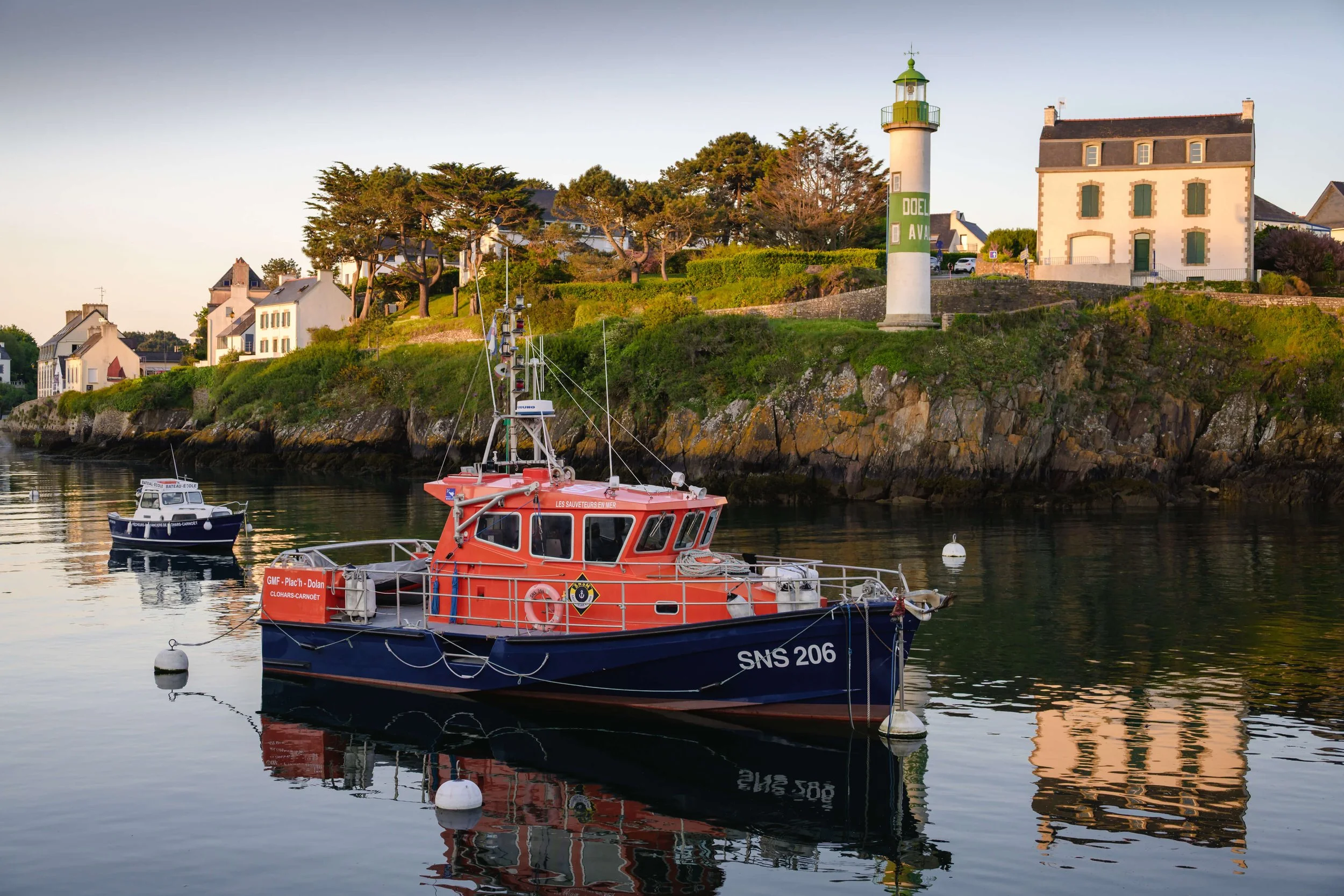 Bateau de sauvetage rouge et bleu amarré dans un port, avec un phare à fond de côte et des maisons blanches au sommet de la falaise