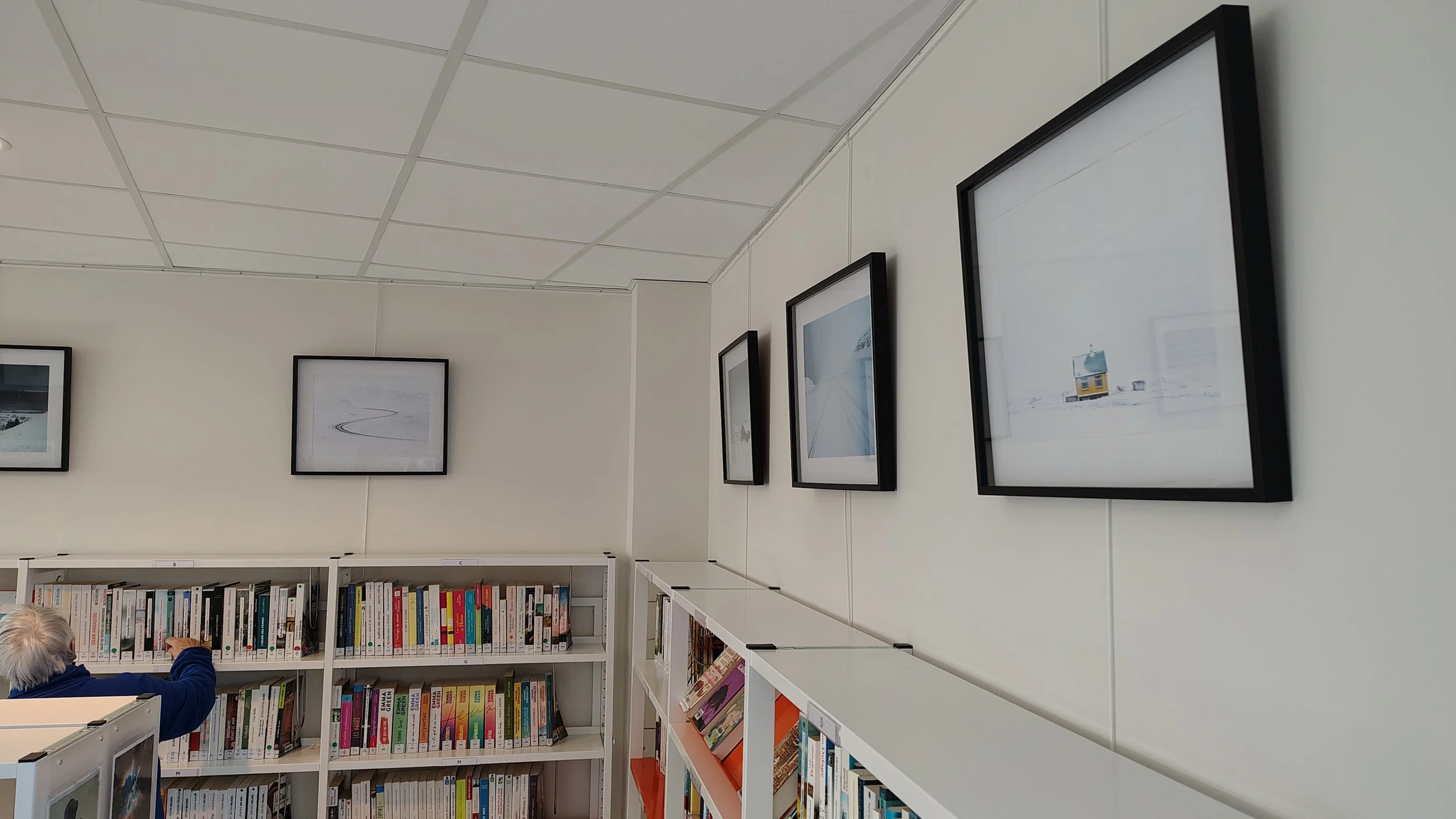Une salle avec des étagères remplies de livres, une personne âgée pointant du doigt vers une étagère, dans une bibliothèque. La murale est décorée avec des photos encadrées de paysages enneigés.