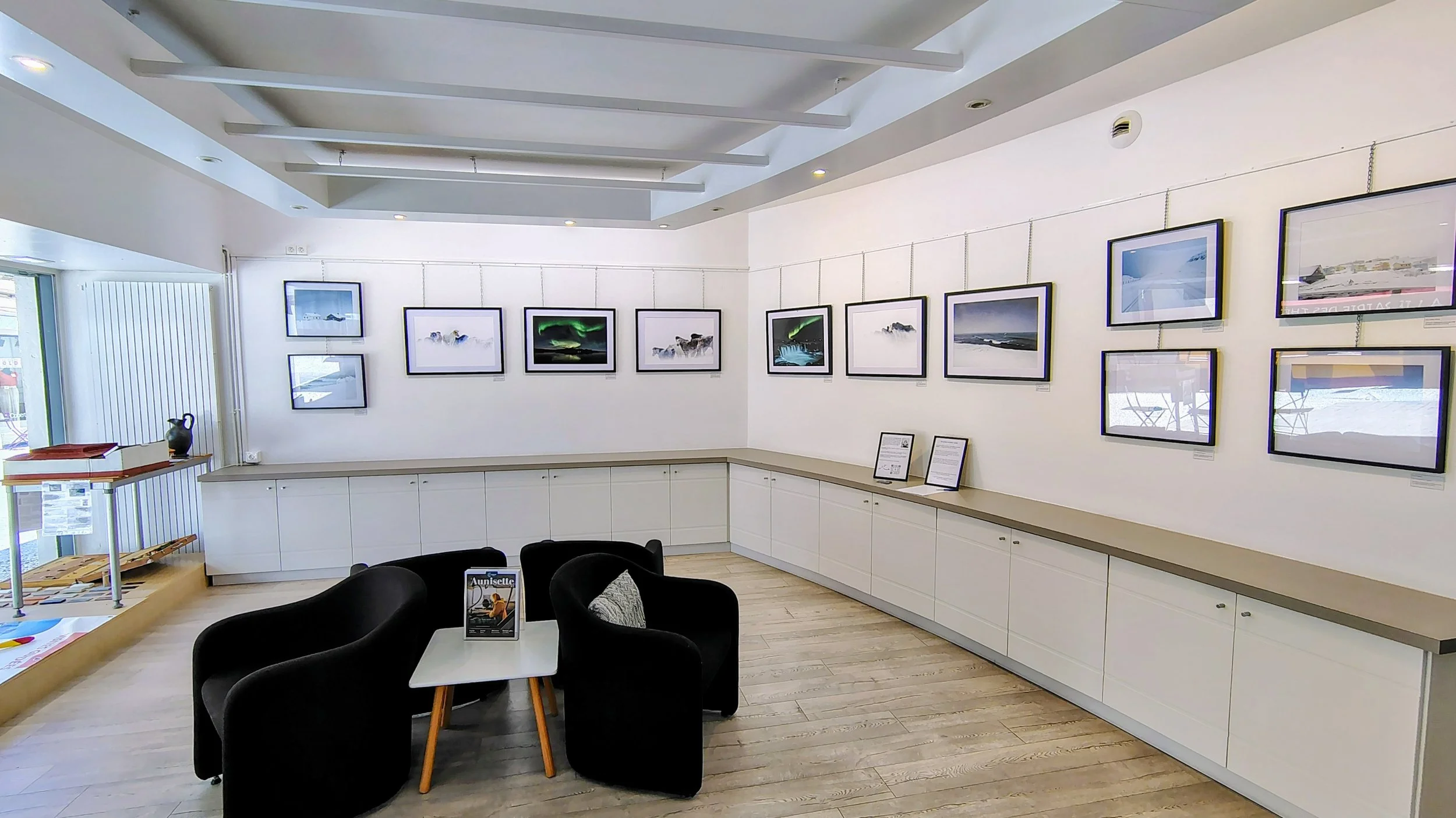 Salle d'exposition avec plusieurs tableaux de paysages et aurore boréale suspendus au mur blanc, coin salon avec quatre fauteuils noirs, table blanche avec magazine, étagère avec objets décoratifs, sol en bois.
