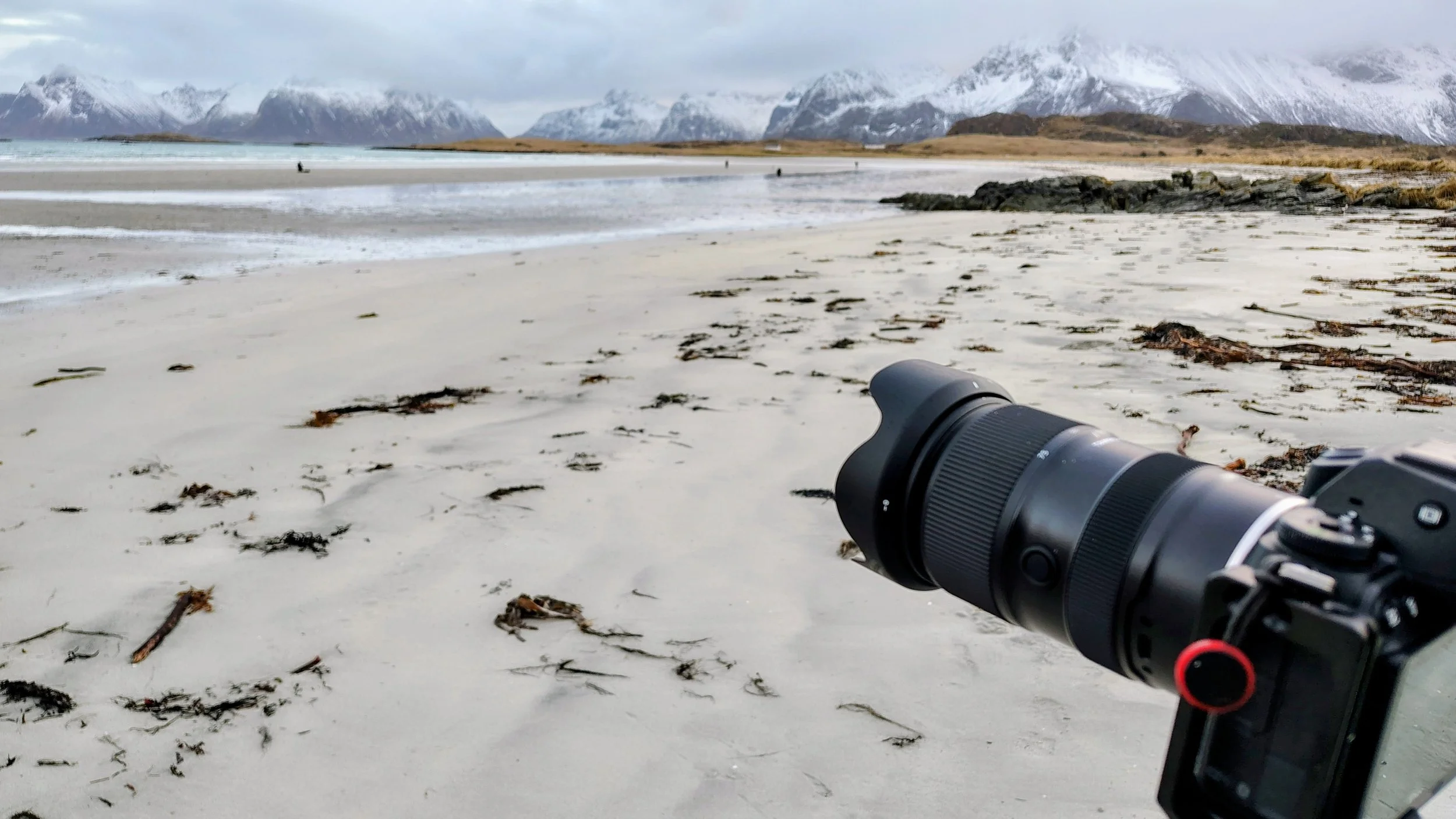 Une plage avec du sable blanc, des rochers, et des montagnes enneigées en arrière-plan. Un appareil photo est visible au premier plan à droite.
