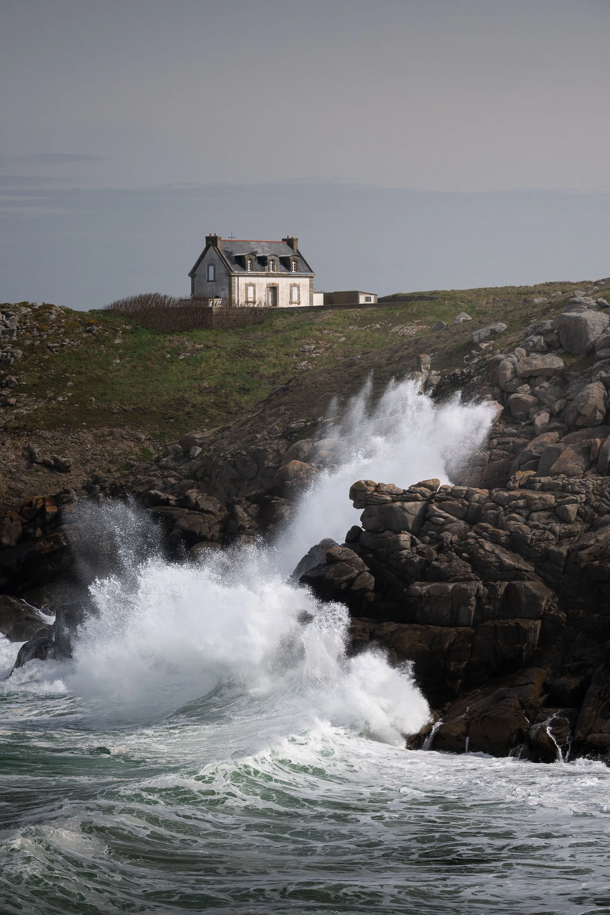 Vagues puissantes se brisant sur une côte rocheuse, avec une maison isolée en arrière-plan sous un ciel couvert.