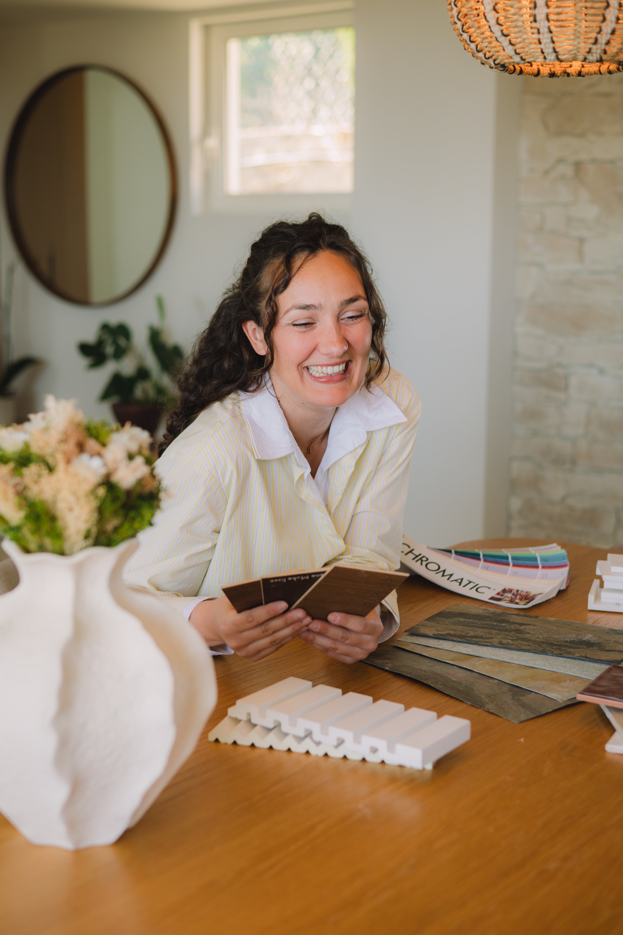 Une femme souriante regarde des échantillons de couleurs, assise à une table où sont disposés des échantillons, des catalogues et des plans de décoration.