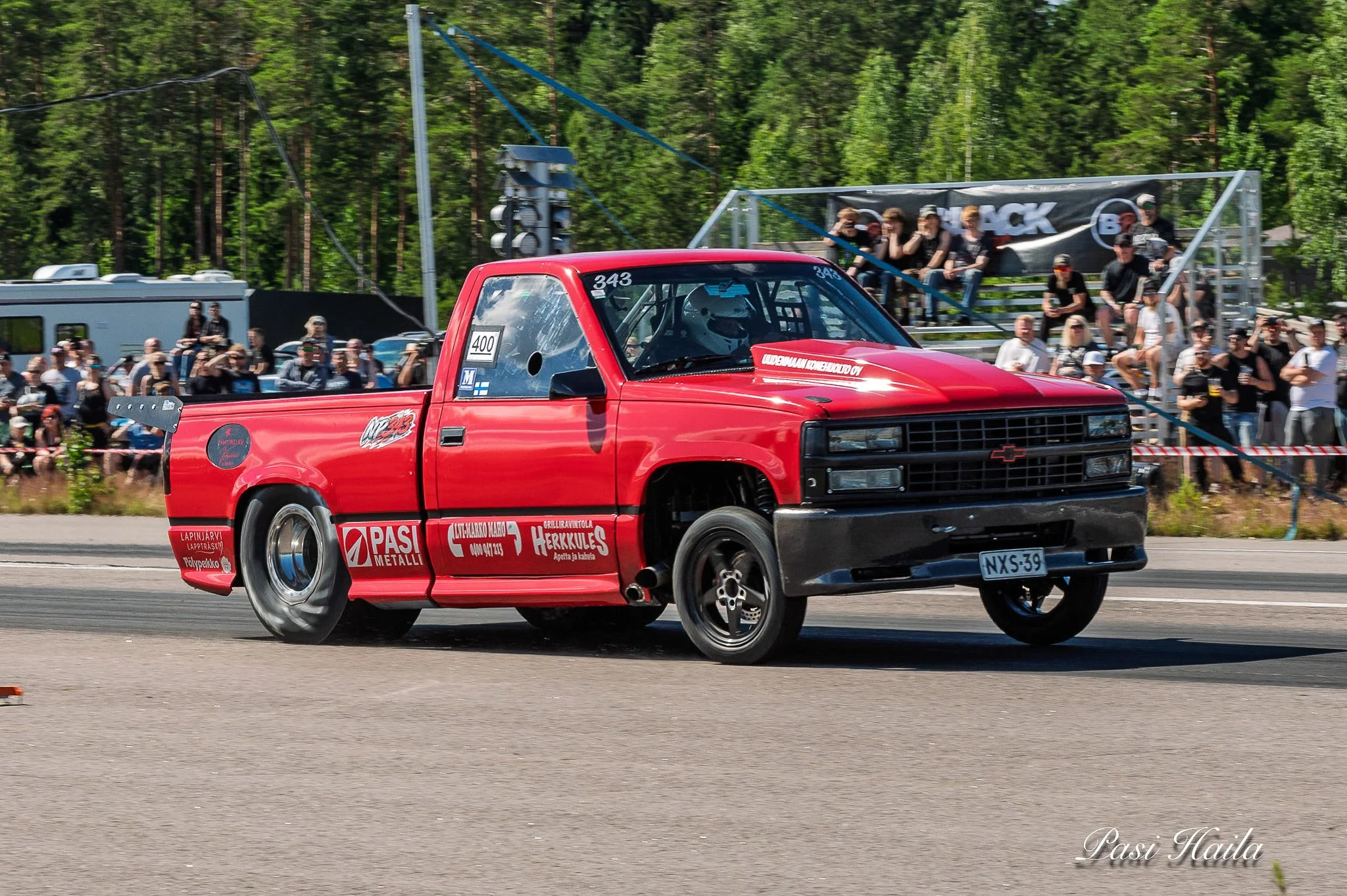 Racecar on track with team members and spectators in background at a racing event.