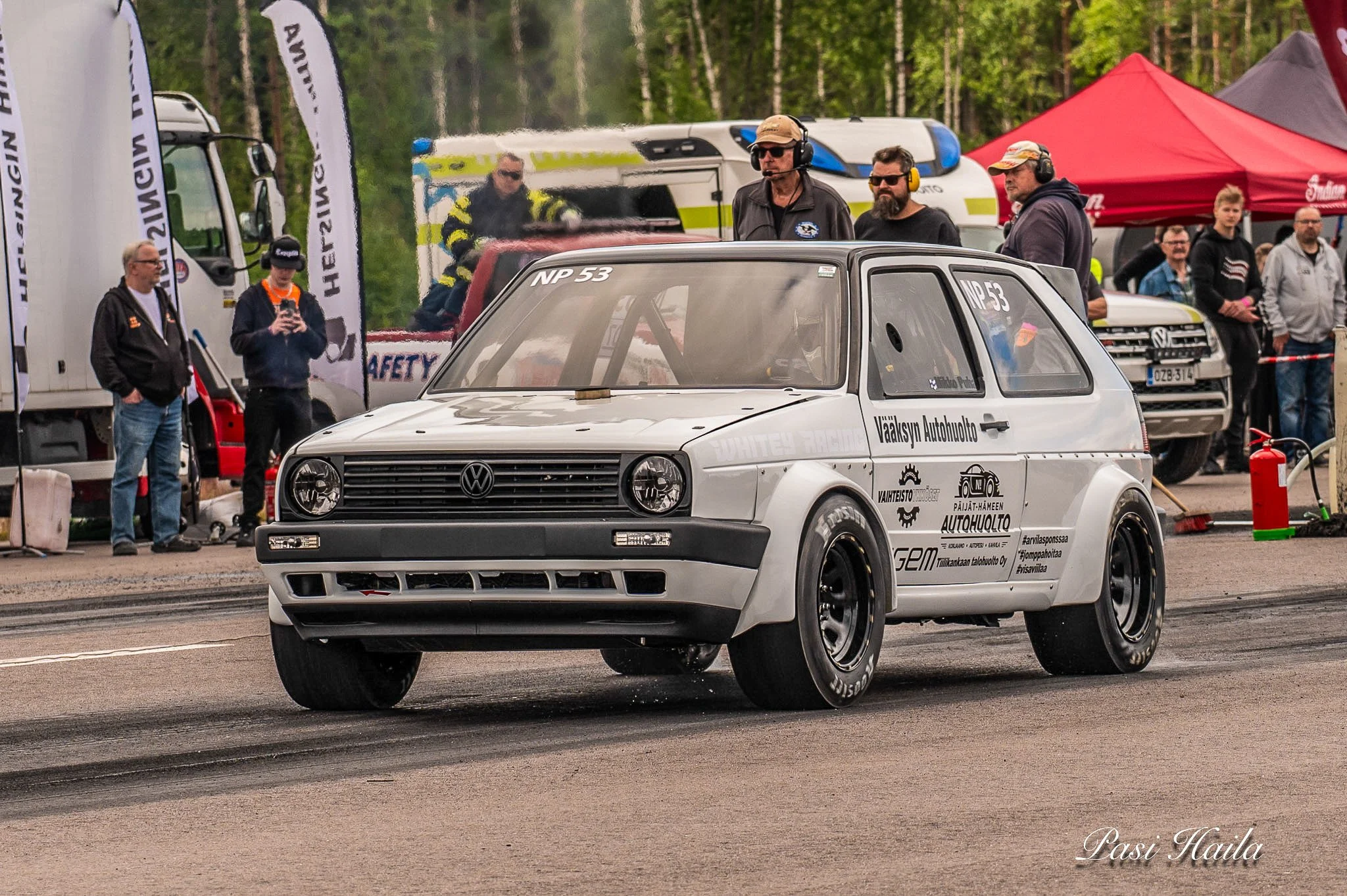 Valkoinen Volkswagen Golf kilpa-auto autoitsemissä tapahtumassa, oikealta eteenpäin ajava, taustalla ihmisjoukkoja ja tiimiä. Auto on varustettu kilpailusettein ja ralleihin tarkoitettuin renkaisiin.