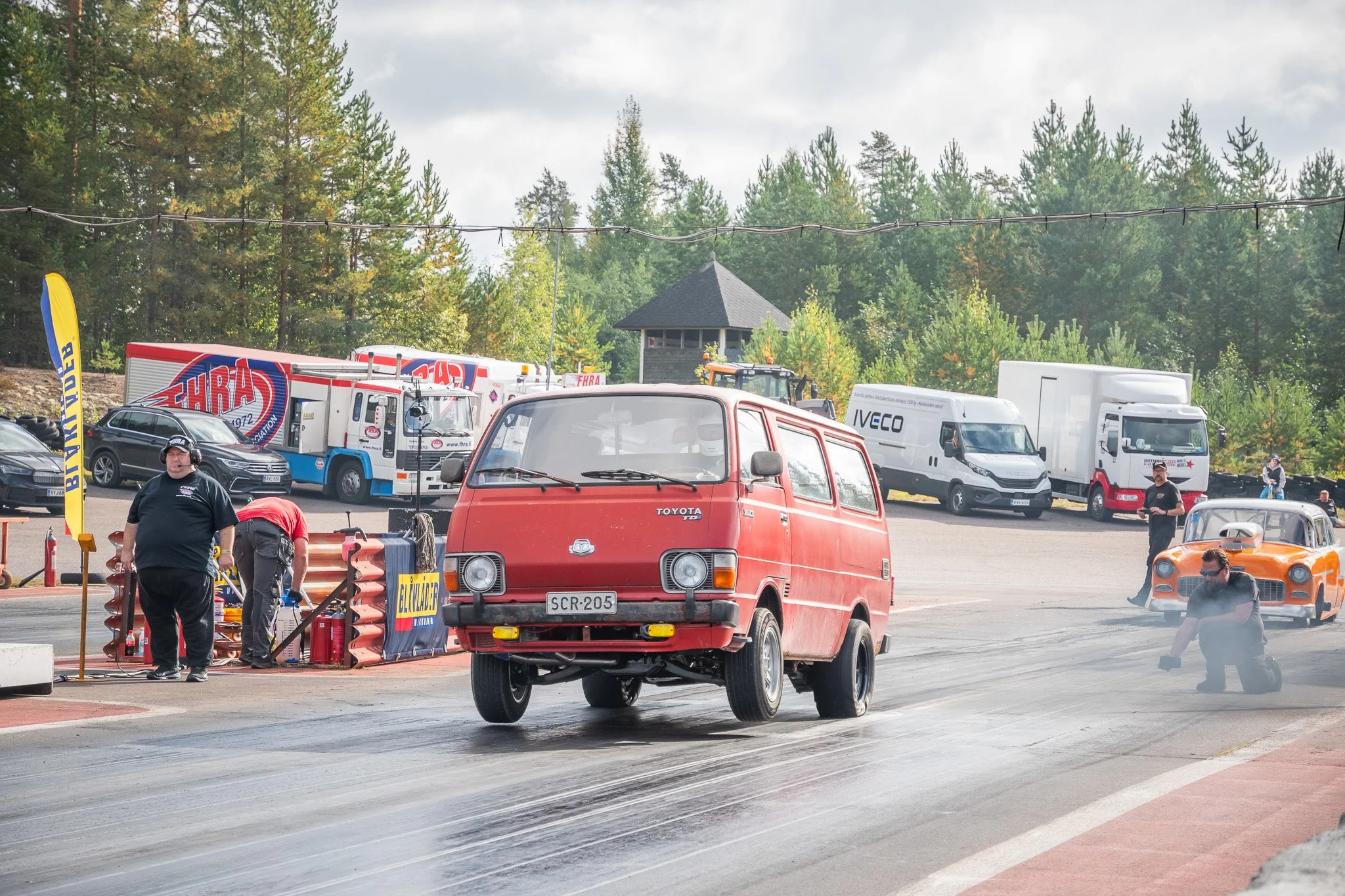 Animeistunut musta klassikkoauto kiihtyy nopeudella, jättäen valojuovia jälkeensä, kilpailurata taustalla.
