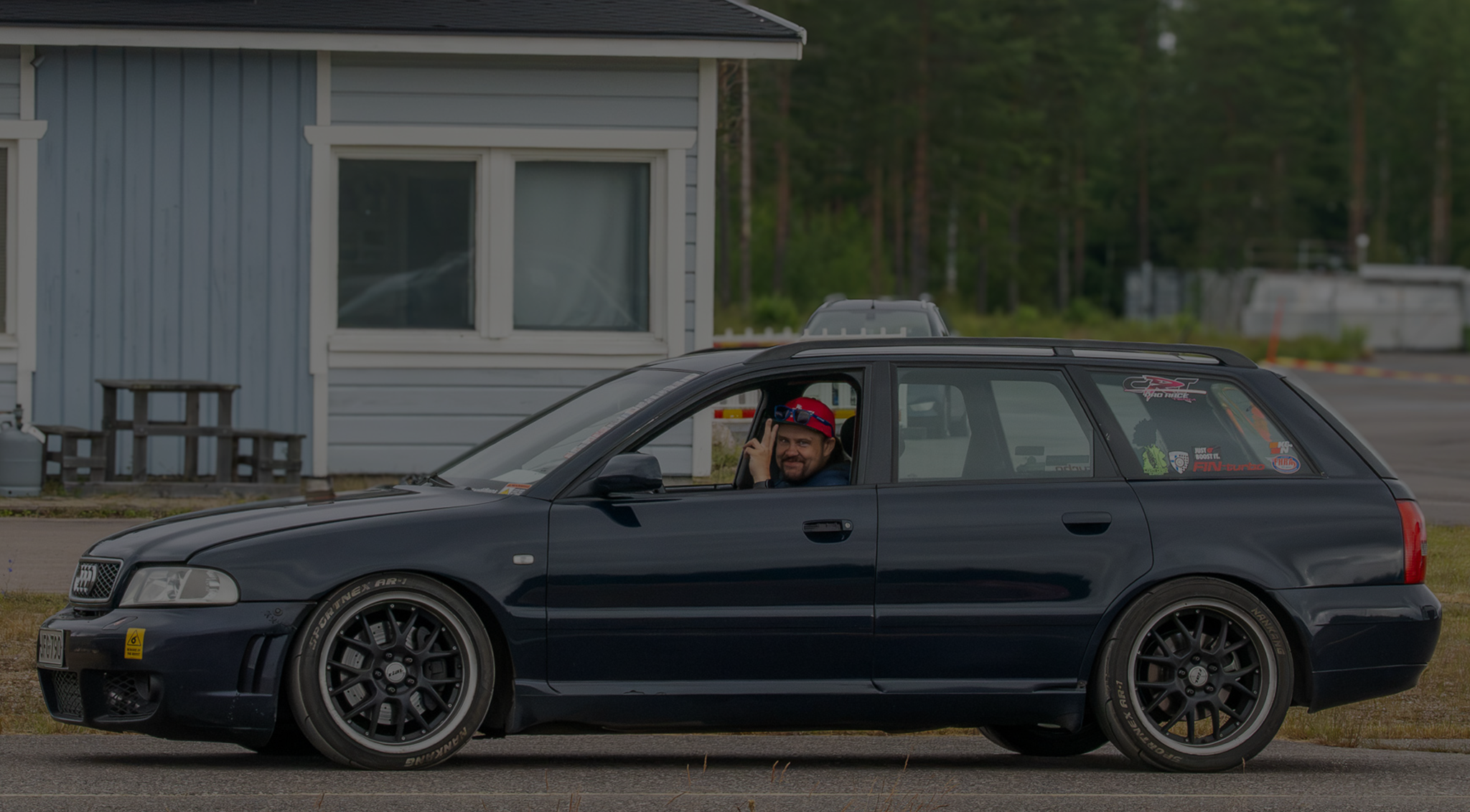 Mustang urheiluauto, joka tuottaa savua, parkissa tai kilpailussa, taustalla muita ajoneuvoja ja rakennuksia, pilvipoutainen taivas.