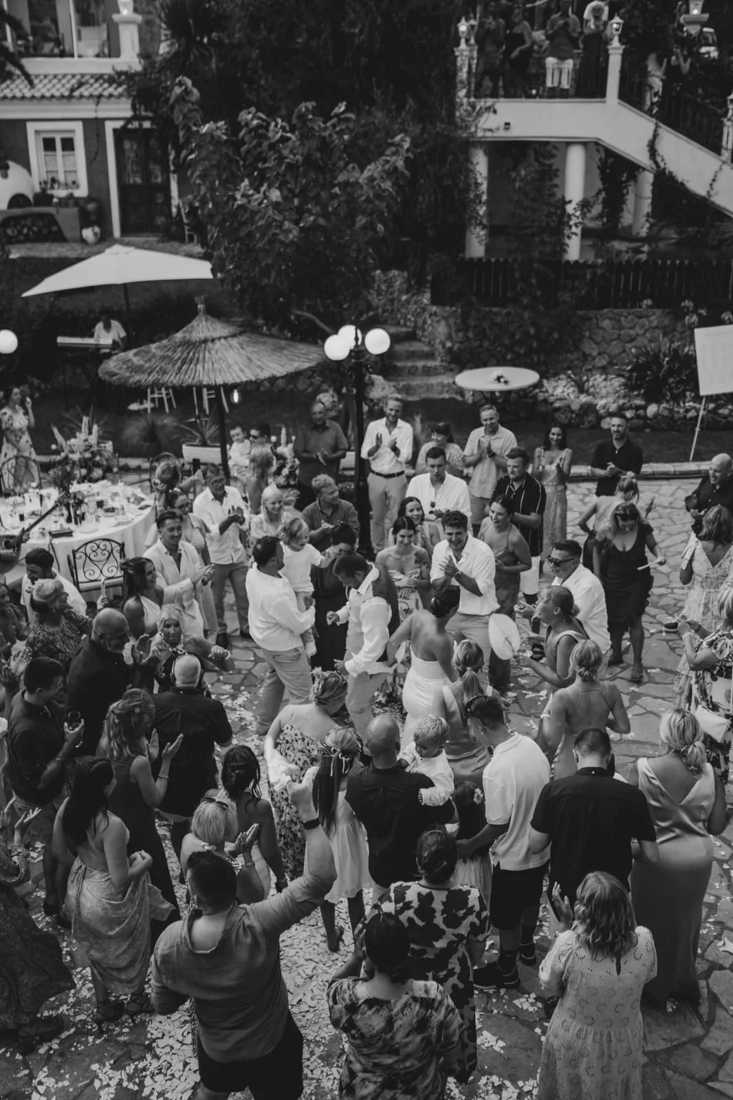 Guests dancing and celebrating outdoors at a wedding reception, with tables, an umbrella, and a thatched roof canopy visible.