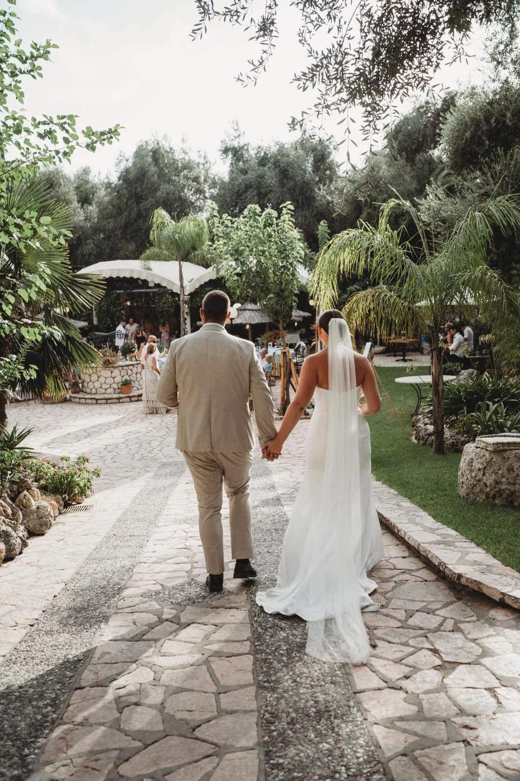 A bride and groom holding hands as they walk down a stone pathway in a lush garden during their wedding celebration.