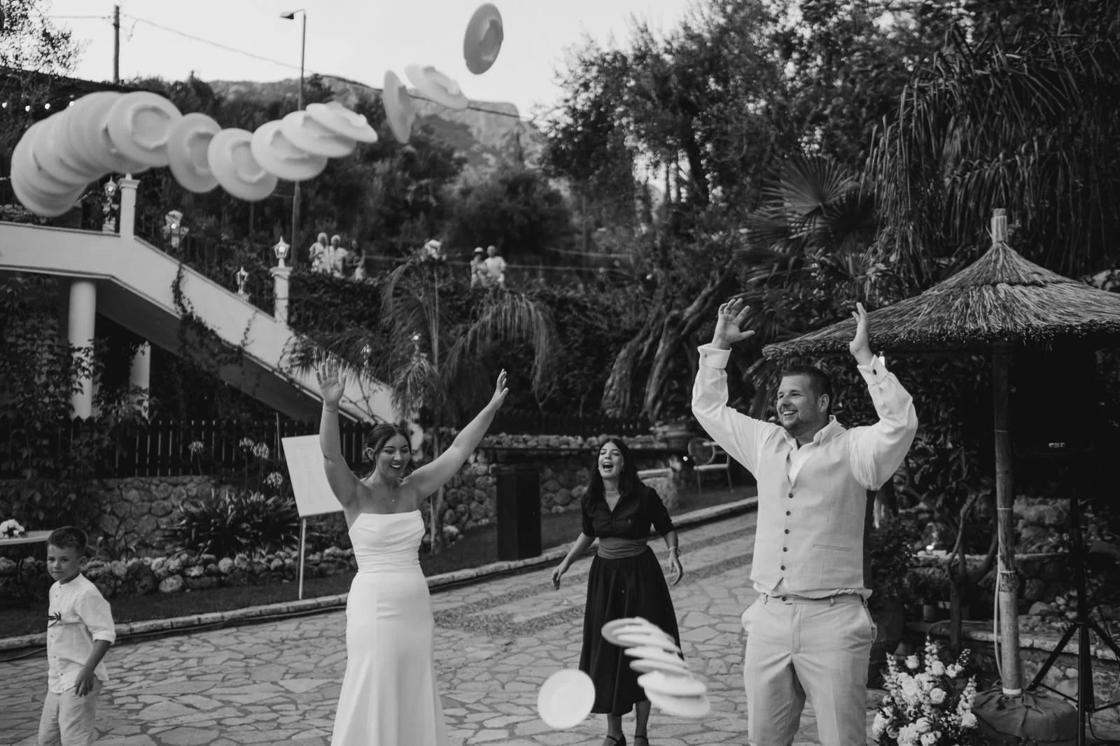 Group of people celebrating outdoors with floating paper plates and a woman in a white dress, likely at a wedding reception.