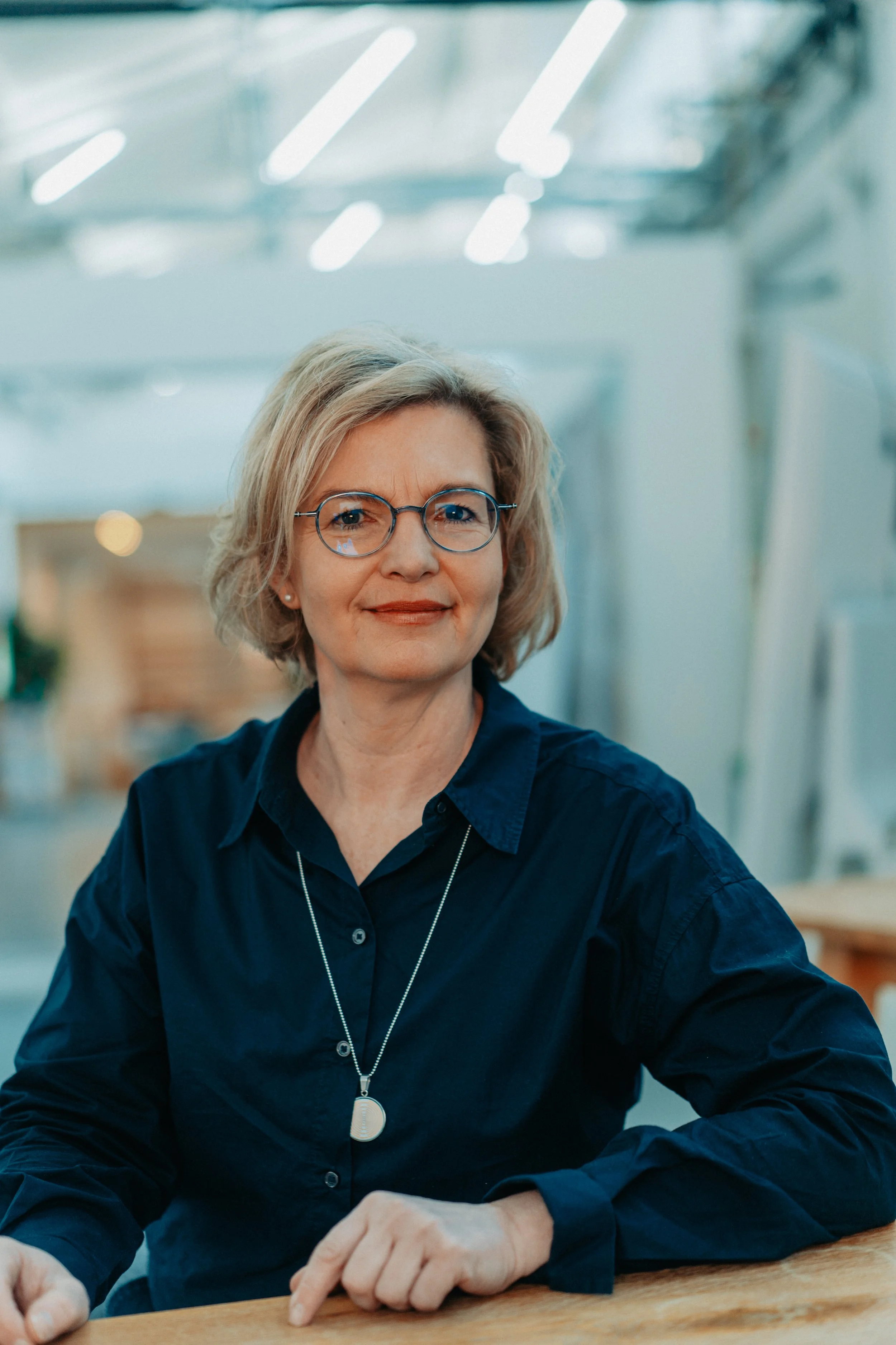 Petra Beuthel. Sie ist eine Frau mit blonden Haaren und einer Brille. Sie trägt eine dunkelblaue Bluse, sitzt an einem Tisch in einem modernen, gut beleuchteten Raum und lächelt leicht.