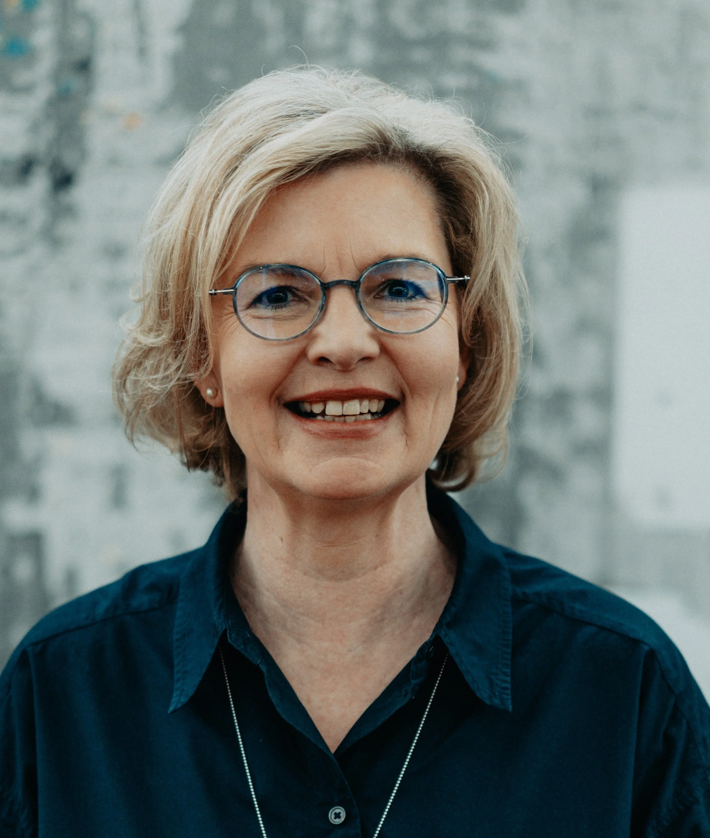 Nahaufnahme von Petra Beuthel. Sie ist eine Frau mit blonden Haaren. Sie lächelt, trägt eine Brille, eine blaue Bluse und eine Halskette.