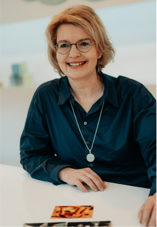 Petra Beuthel. Sie ist eine Frau mit blonden Haaren. Brille und einer blauen Bluse. Sie lächelt und sitzt an einem Tisch. Auf dem Tisch liegen Karten.