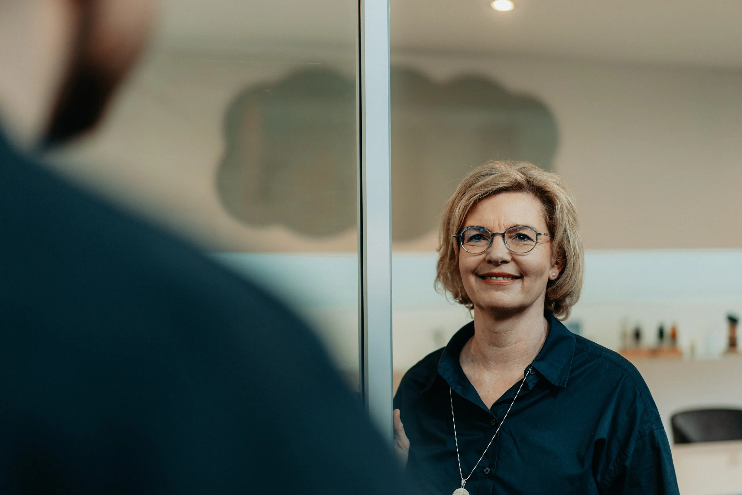 Petra Beuthel. Sie ist eine Frau mit blonden Haaren und Brille. Lächelnd steht sie an einer Glastür und begrüßt jemanden.