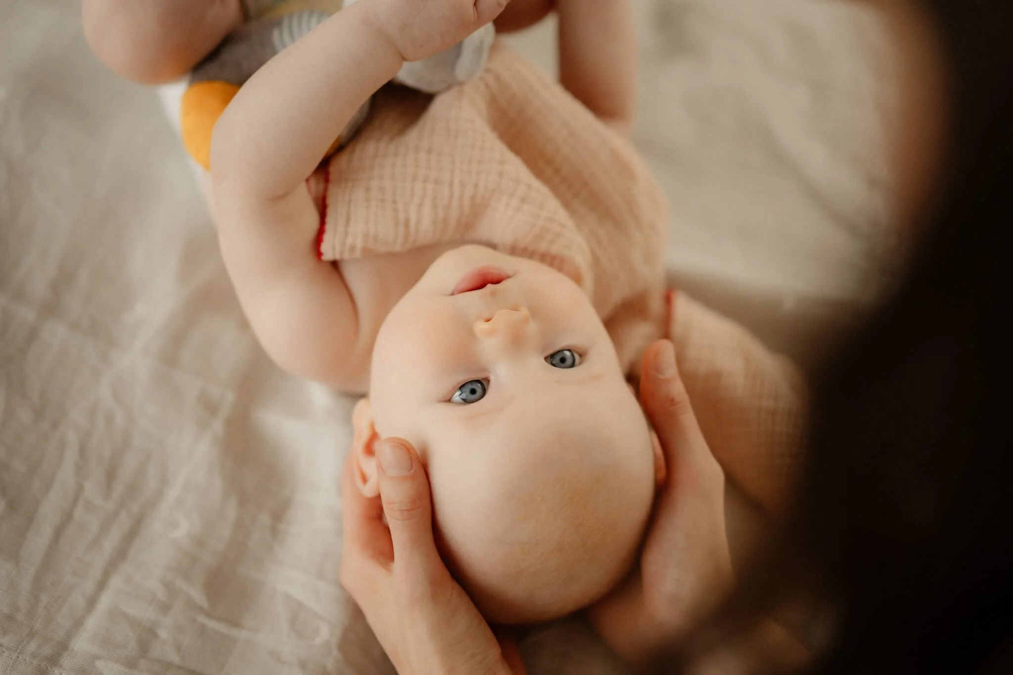 Ein Baby liegt auf einer Decke und wird von einer Person während der Babymassage an den Seiten des Kopfes sanft gehalten. Das Baby trägt einen cremefarbenen Strampler und hat blaue Augen.