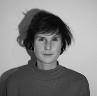 Schwarz-weiß Porträt einer Yoga Lehrerein mit kurzem Haar und Ohrringen, die direkt in die Kamera schaut, vor einem neutralen Hintergrund.