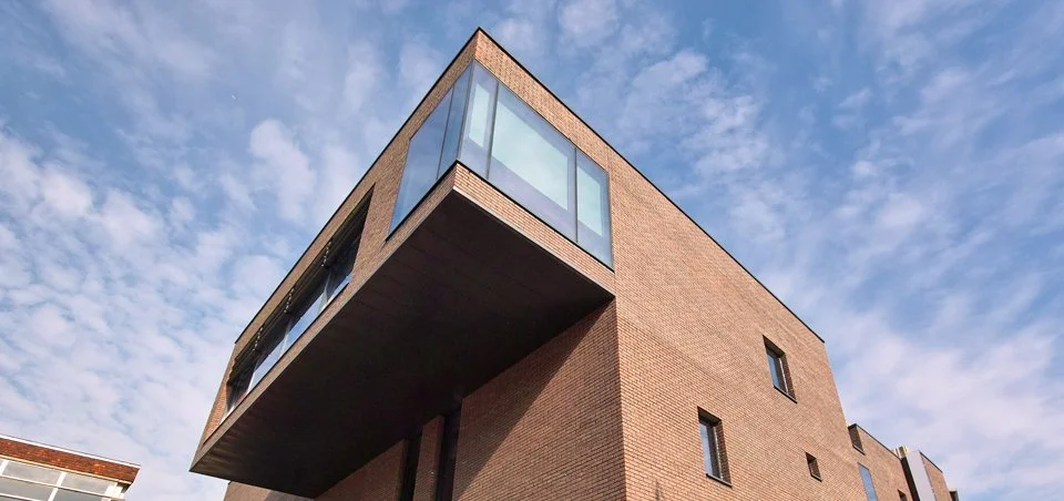 view up to underside of cantlivered room. Blue sky, brown brickwork, reflective windows.