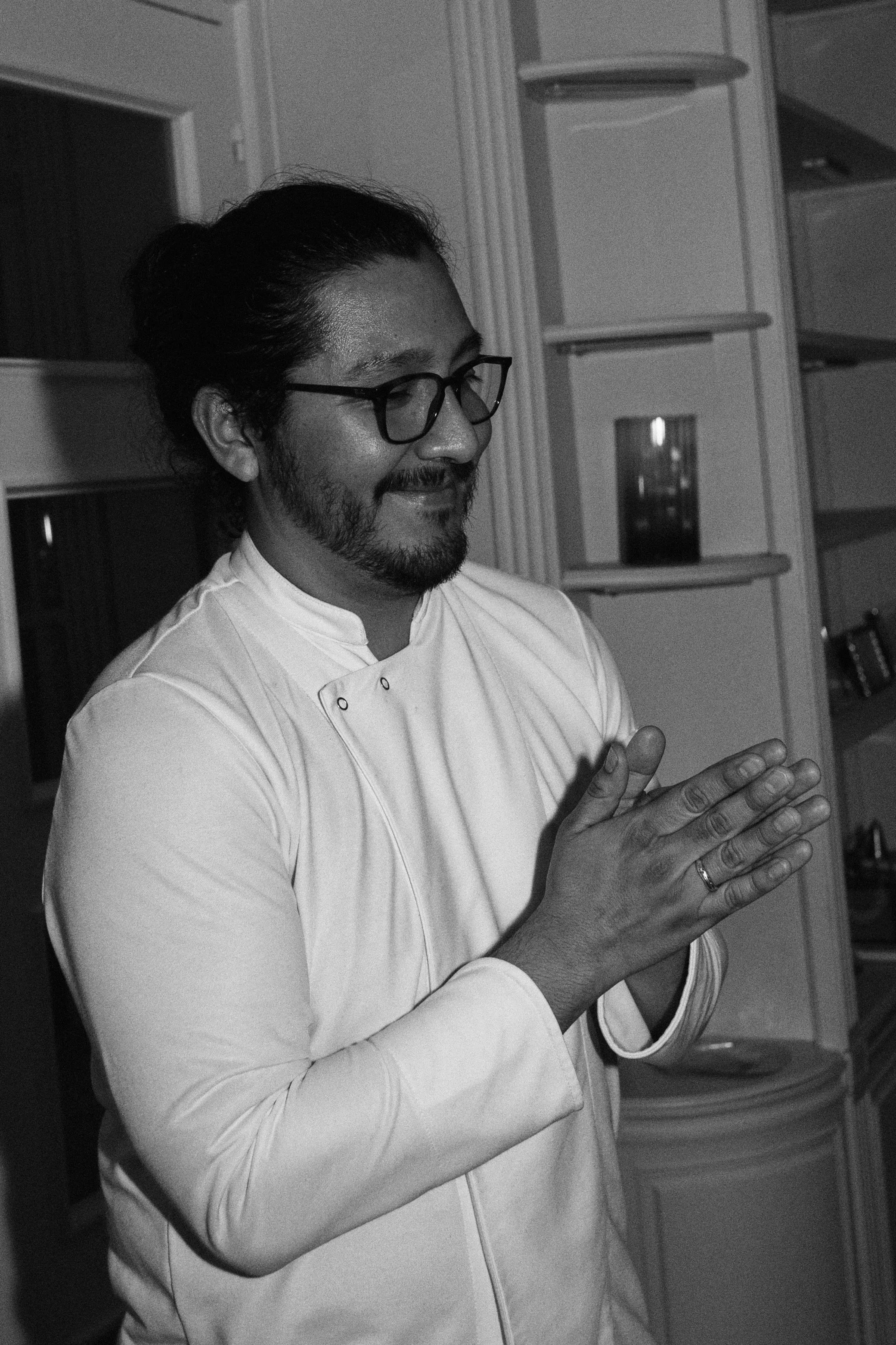A man with glasses and a beard, wearing a chef's coat, is smiling with his eyes closed and hands clasped in front of him, indoors with shelves in the background.