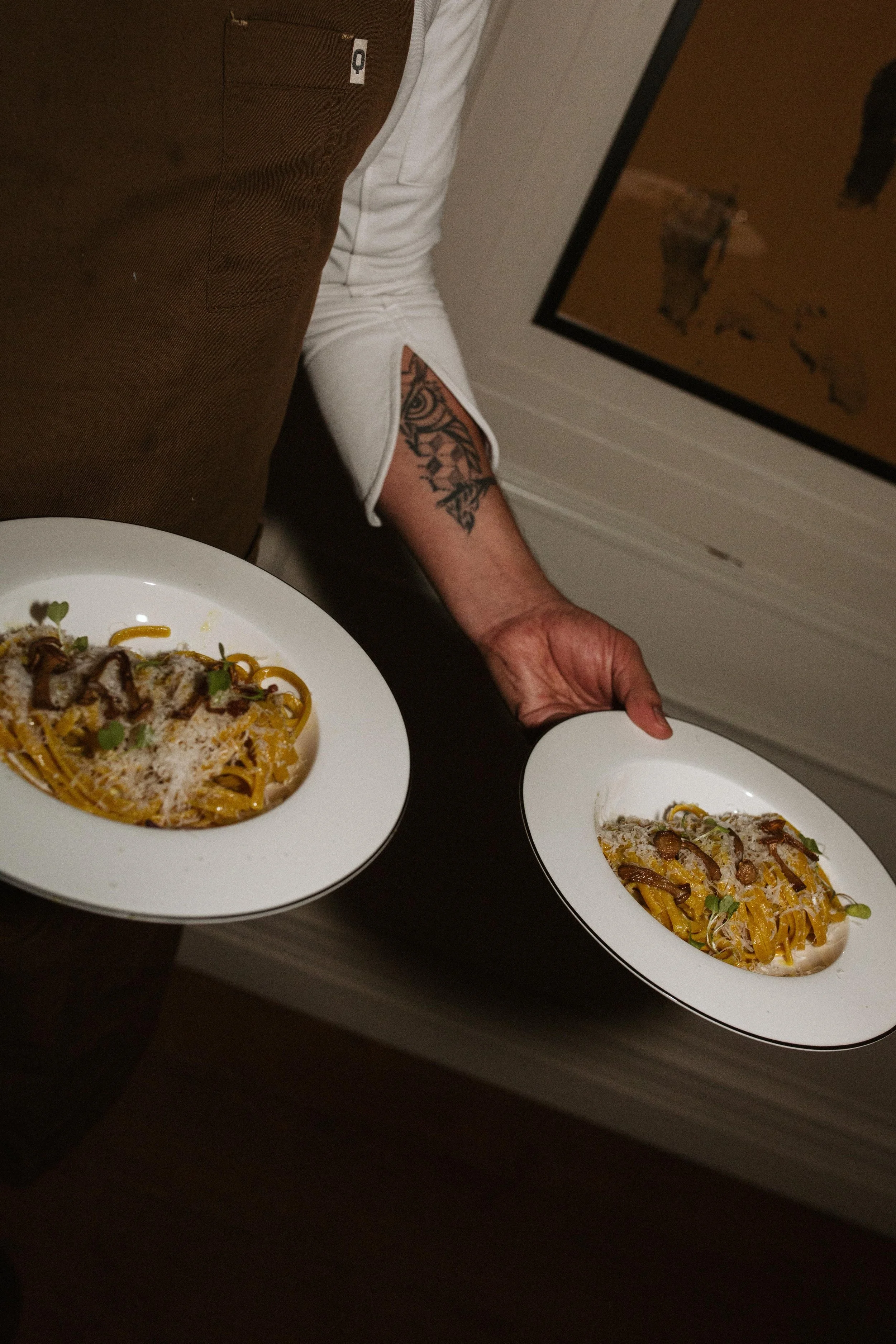 Two plates of fettuccine pasta with sauce, cheese, and garnishes, held by a person wearing a white rolled-up sleeve, standing near a mirror and wooden door. Meals crafted with care and intention.
