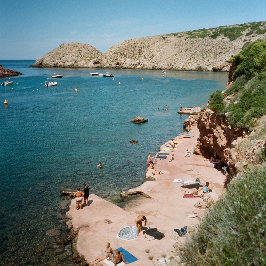 A scenic coastal scene with a rocky beach, people sunbathing and swimming, and boats anchored in a calm bay surrounded by cliffs and greenery. Menus rooted in seasonality.