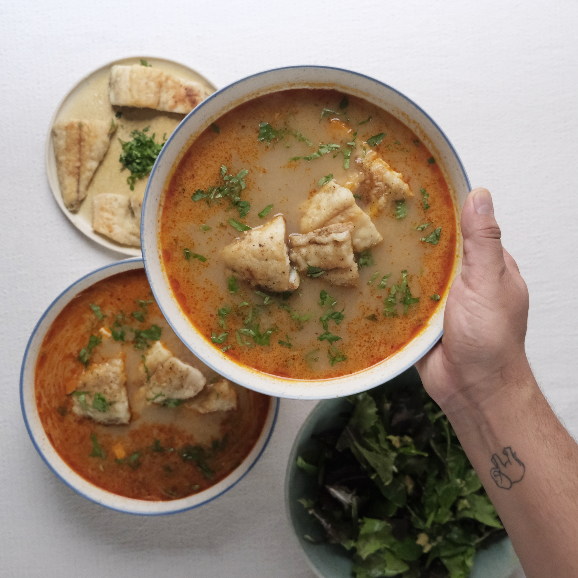 A hand holding a bowl of creamy soup garnished with chopped herbs, with a plate of bread and greens in the background.