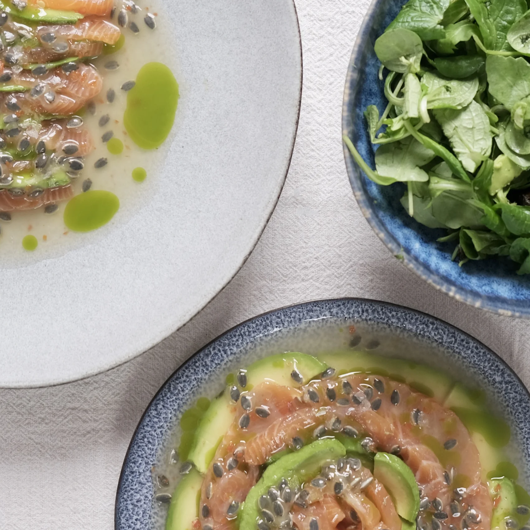Close-up of two bowls of salmon ceviche topped with black seeds, served with avocado slices, and a salad of mixed greens in a blue bowl, all on a light-colored table.