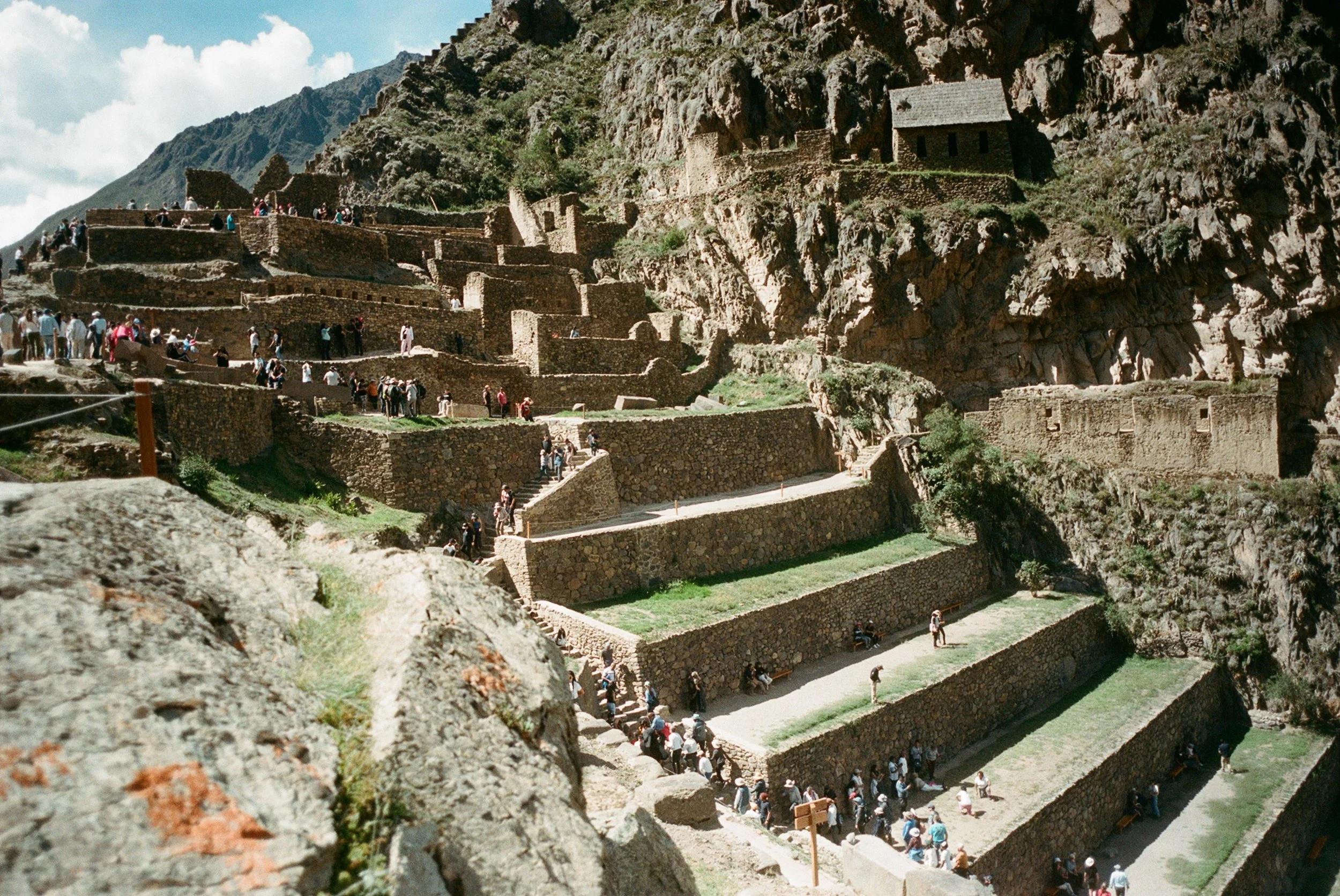 Terraced archaeological site with stone retaining walls and staircases, located on a mountainside with visitors exploring and walking around.