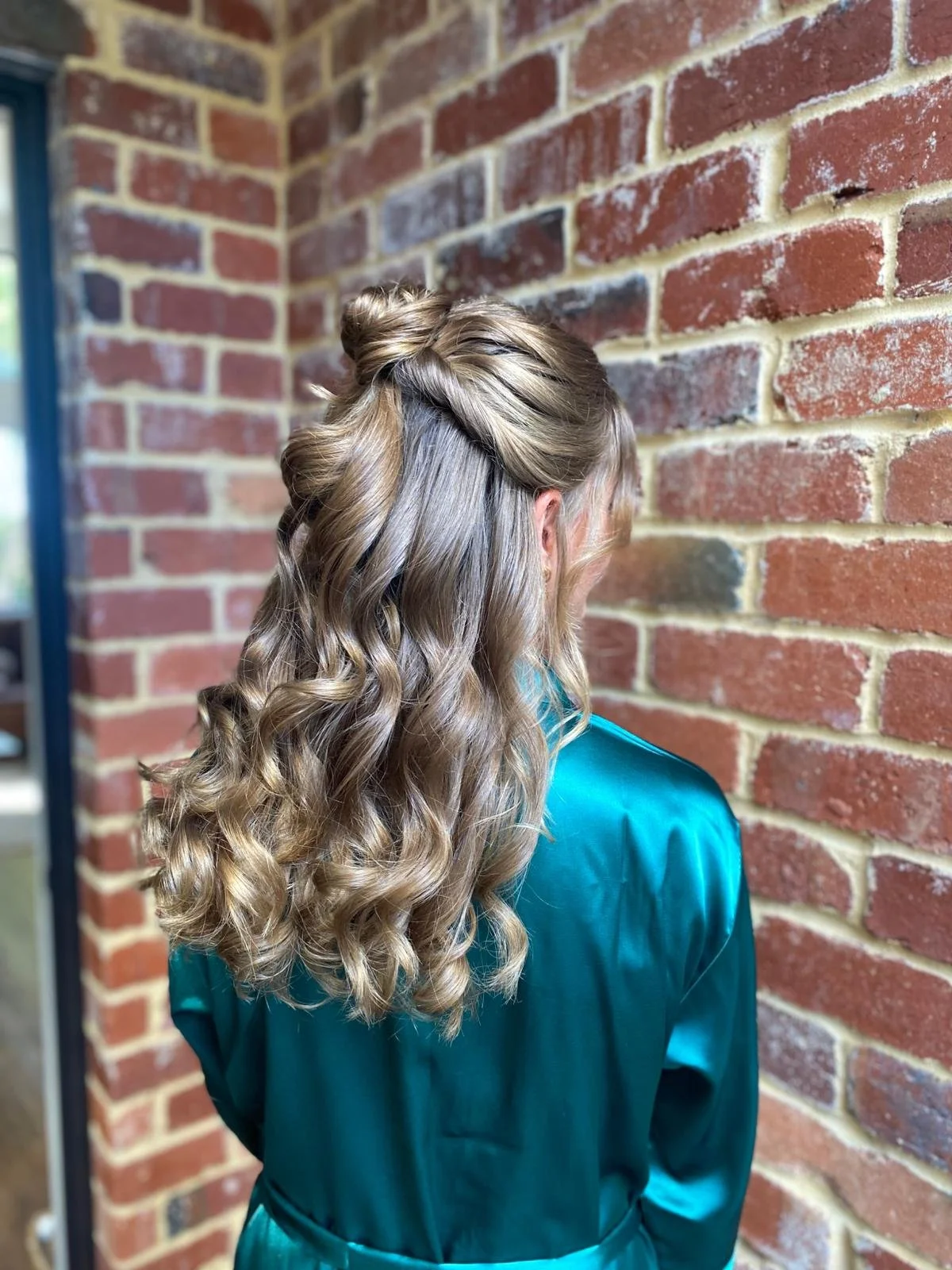 Back view of a woman with long, curled blonde hair styled with half-updo, wearing a teal satin robe, standing against a red brick wall.