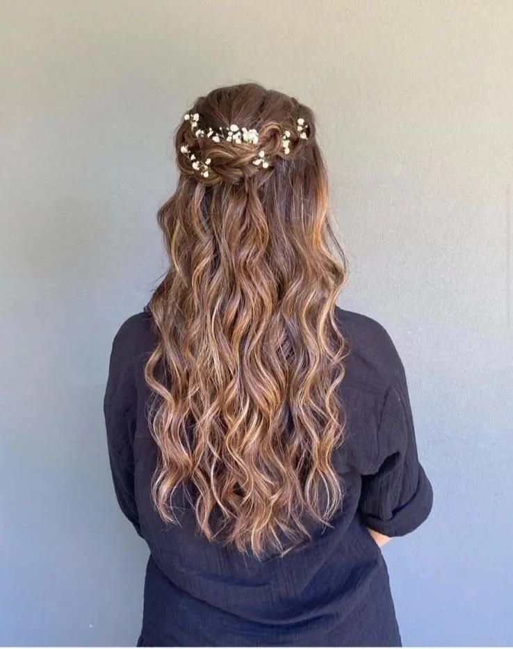 Back view of a woman with long, curly hair adorned with a floral headband, wearing a black shirt, standing against a plain gray wall.