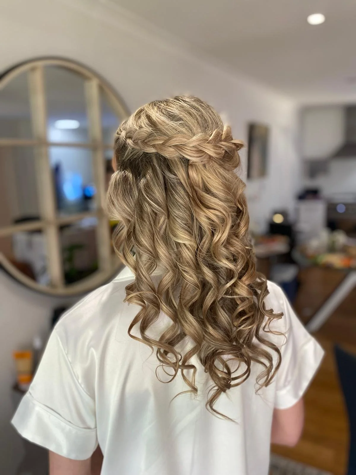 Back view of a woman with blonde, curly hair styled with a side braid across her head, wearing a white top, in a well-lit room.
