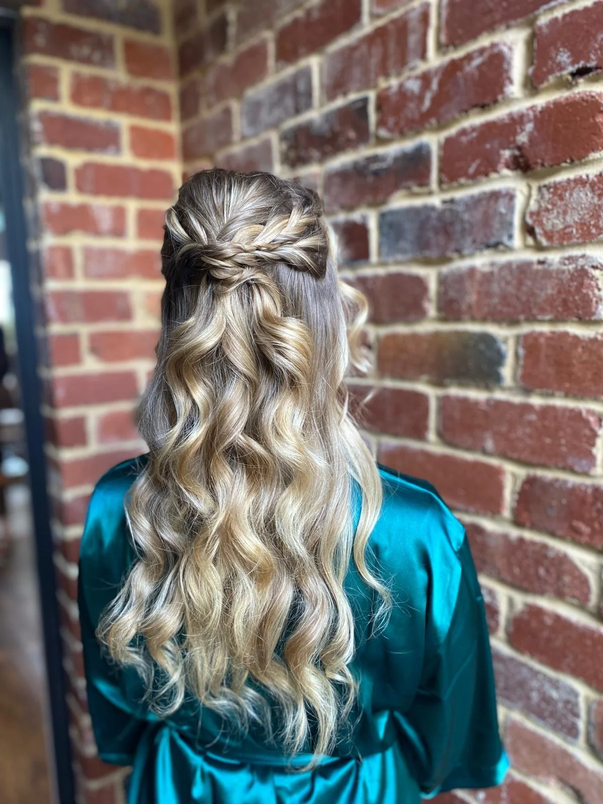 Back of a woman with styled blonde, wavy hair with a twisted braid accent, wearing a teal satin robe, standing against a red brick wall.