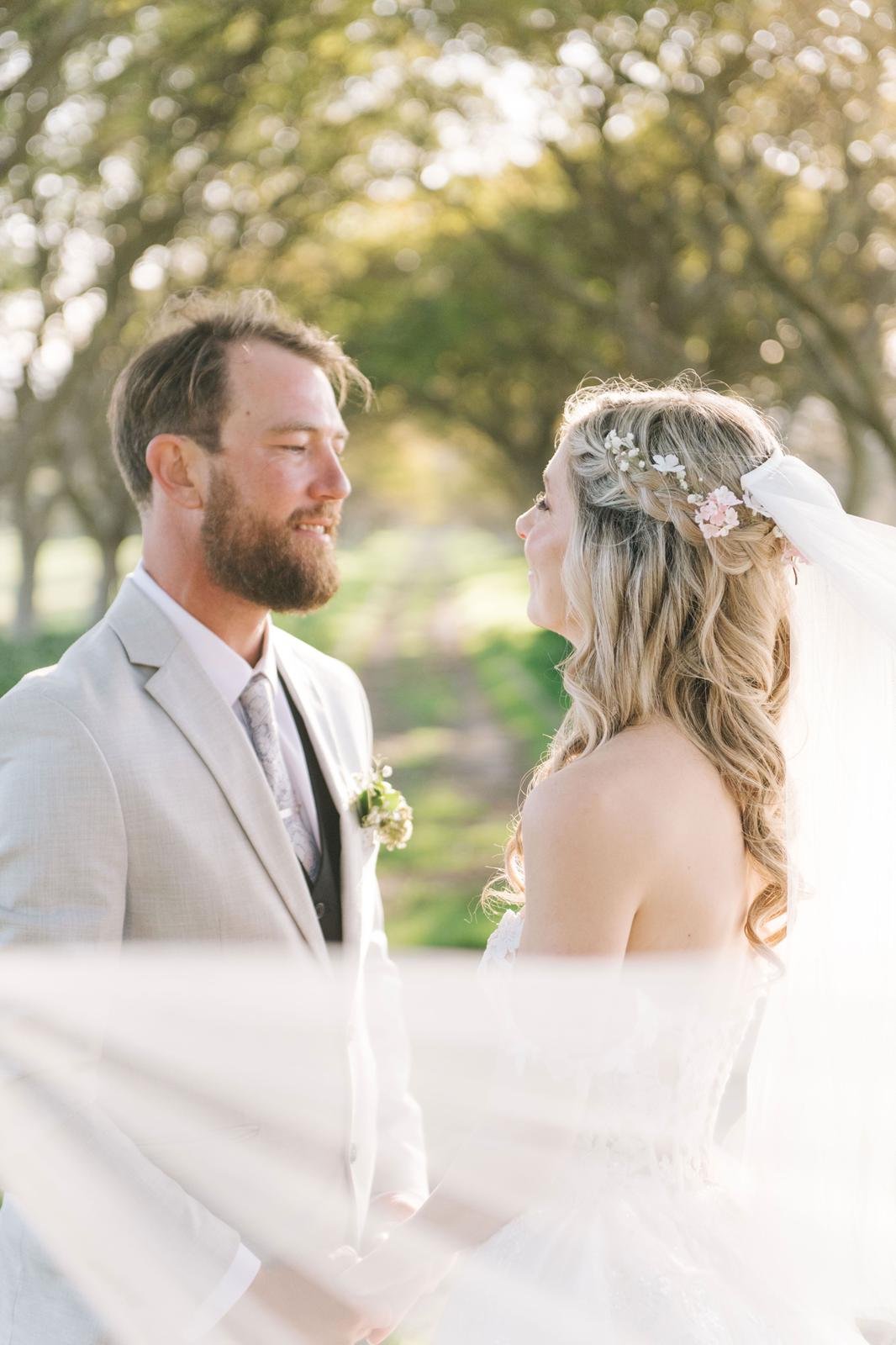 A bride and groom standing close together outdoors. The bride has long, wavy blonde hair with flowers and a white veil. The groom has a beard, short hair, and is wearing a light-colored suit. The setting is a green, tree-lined area with sunlight filt