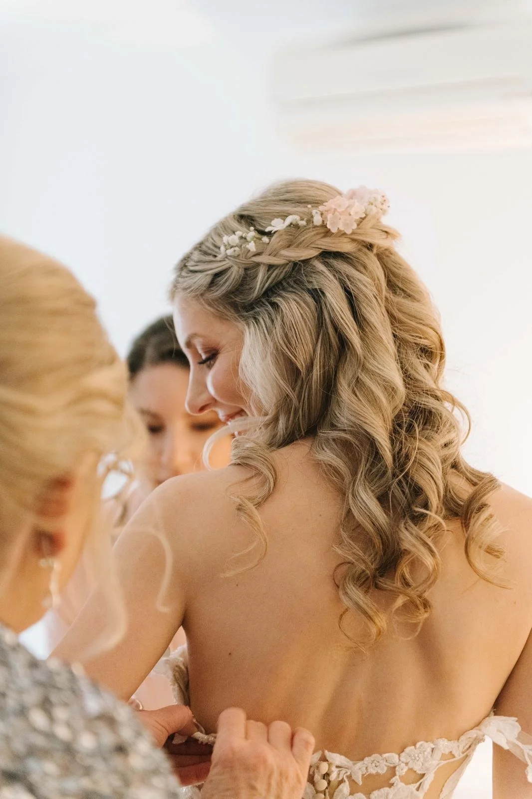 A bride with blonde, curly hair and a floral headpiece is getting dressed by a woman, likely helping her with her wedding dress, while other women look on.