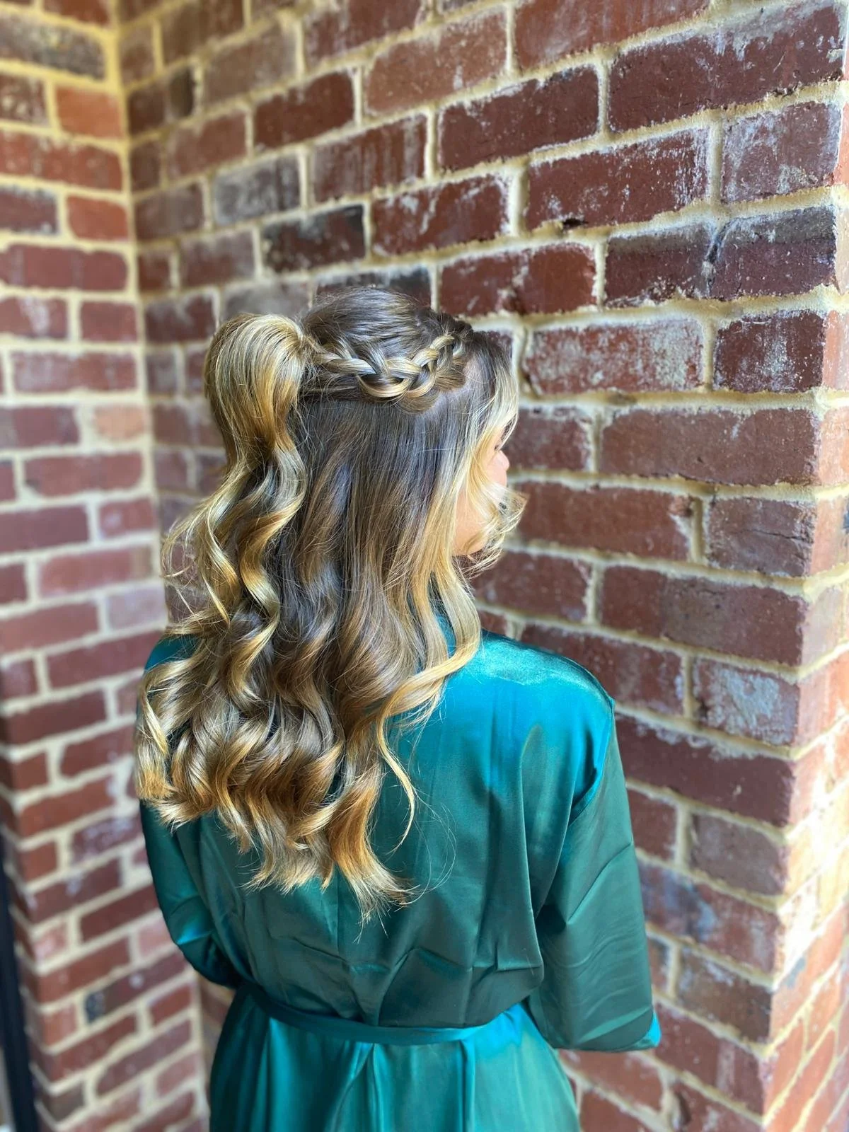 Back view of a woman with long, wavy blonde hair, styled with a crown braid, wearing a teal satin robe, standing against a brick wall.