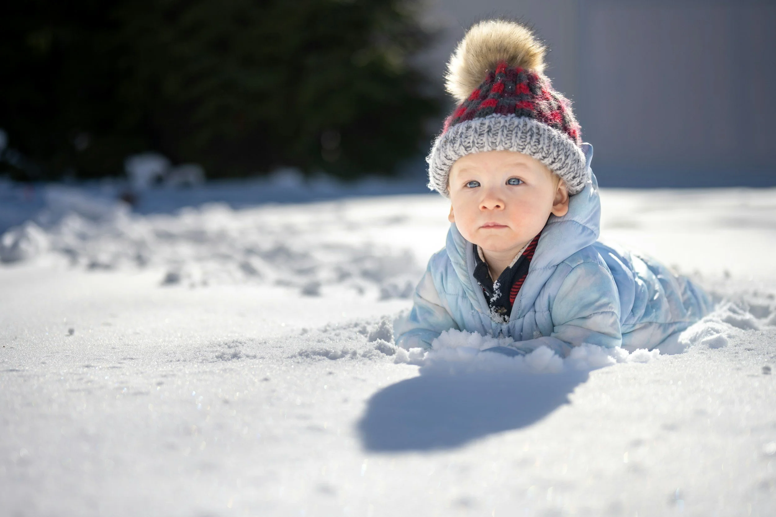 A young child in a blue coat and a knit hat with a pom-pom, lying on their stomach in the snow outdoors with a shadow cast on the snow, in a wintery setting.