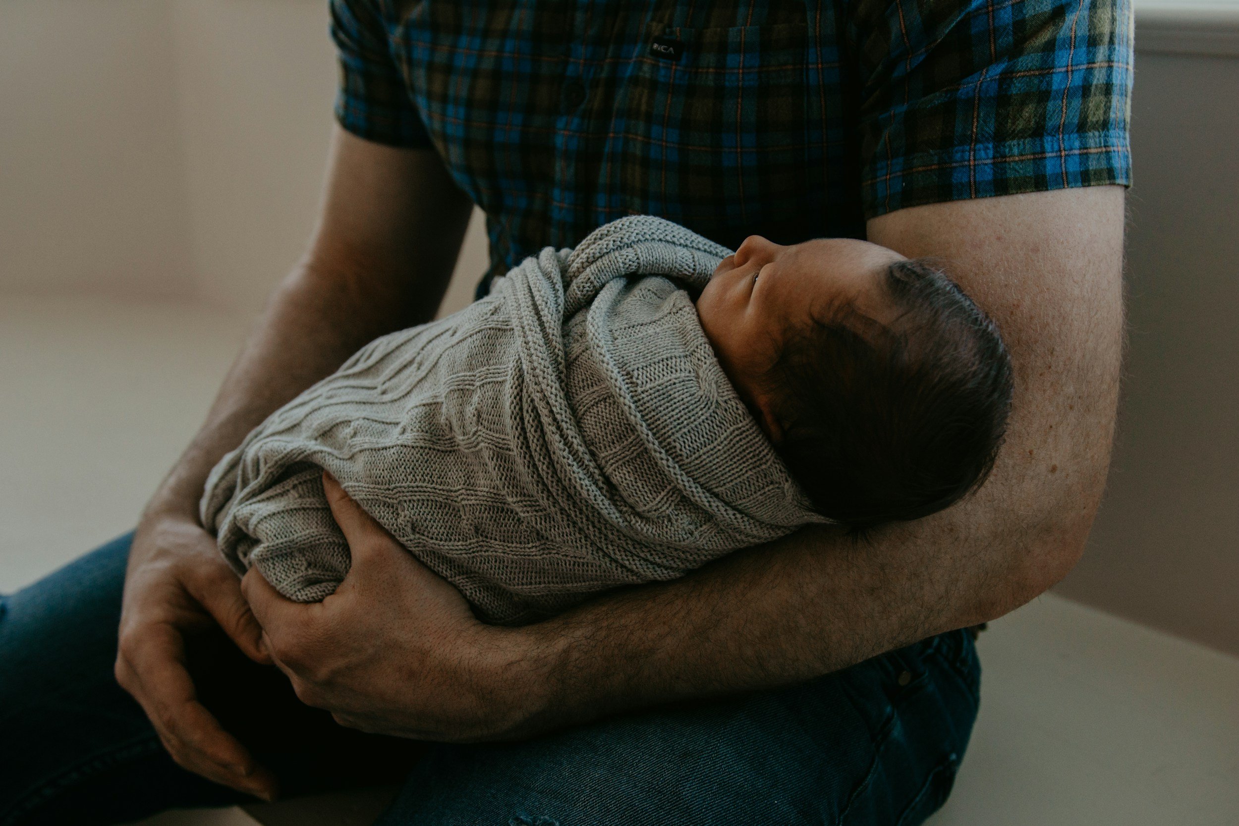 colour photo of a father tenderly holding a newborn baby wrapped in a textured beige blanket, with  a gentle, loving pose.