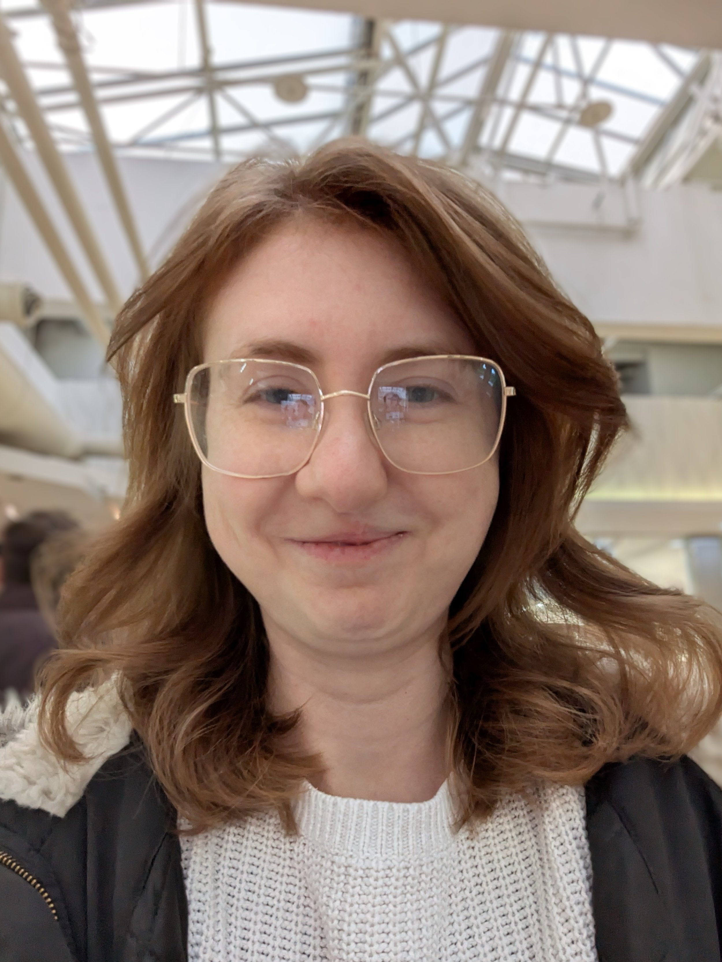 A woman with wavy reddish-brown hair and glasses taking a selfie inside a building with large glass skylights.