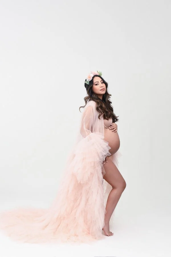 pregnancy image of beautiful long brown hair woman in a pink tulle gown and flower crown. Has both hands on belly on a light background