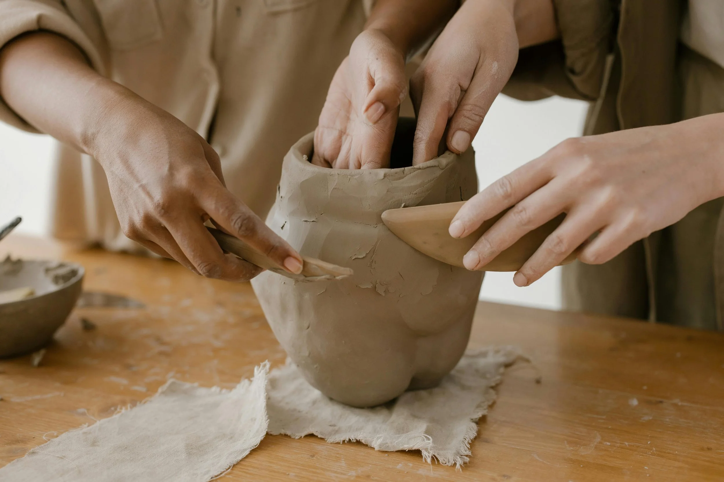 People shaping clay pottery on a wooden table using hands and tools.