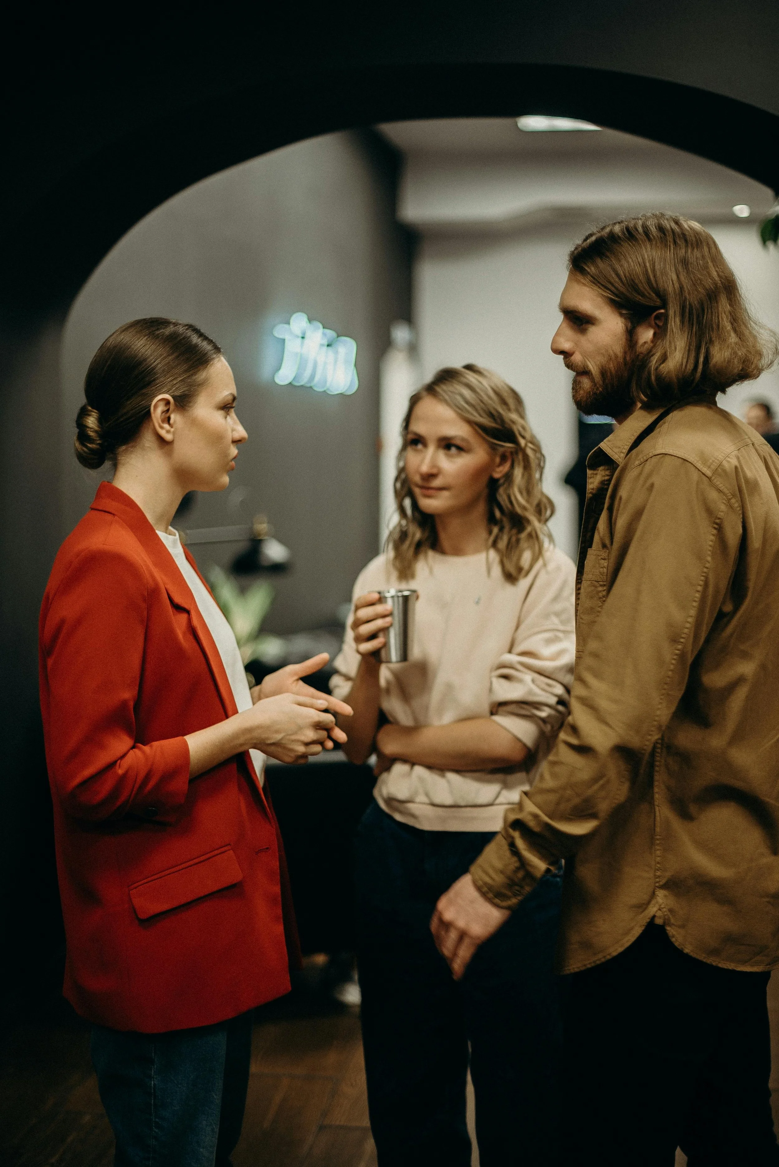 Three people having a conversation indoors, one woman in a red blazer, another holding a cup, and a man wearing a brown shirt.
