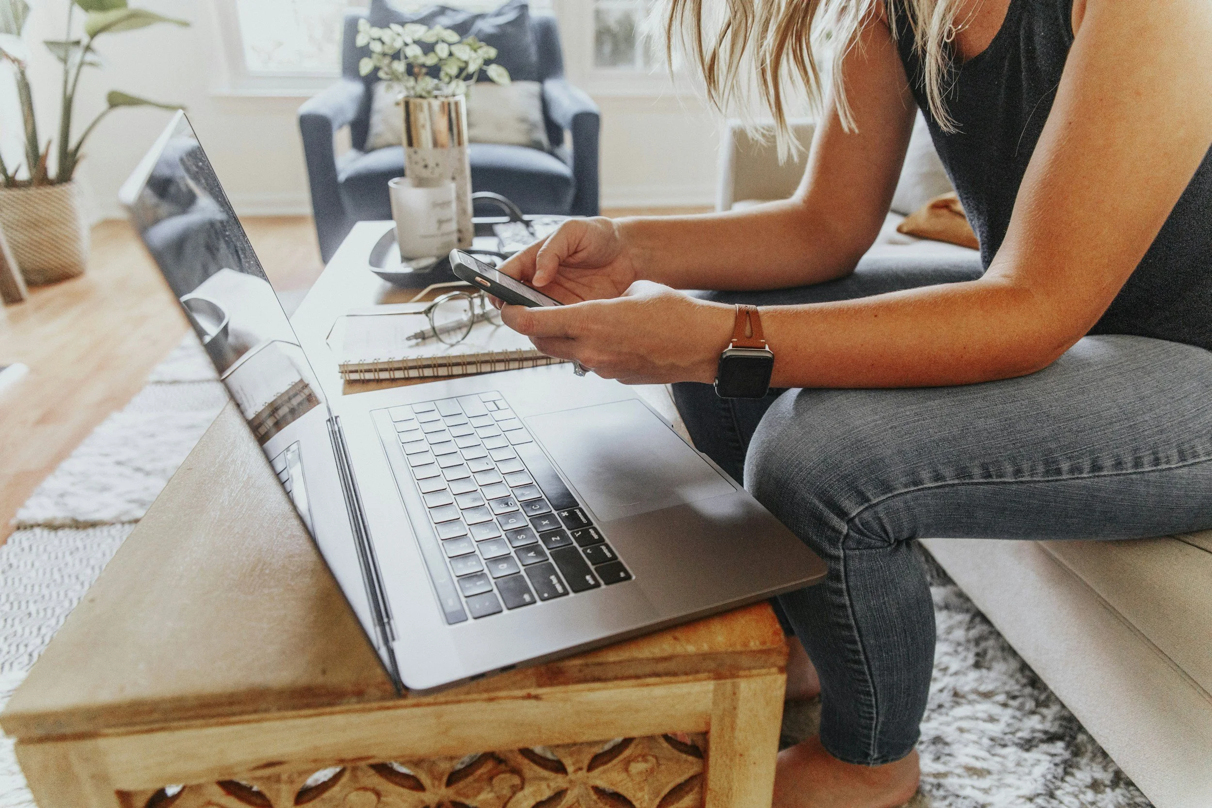 A woman seated on a gray chair, working at a wooden coffee table with a laptop, notebook, glasses, and a smartphone. She is wearing a black sleeveless top, a watch, and a bracelet.