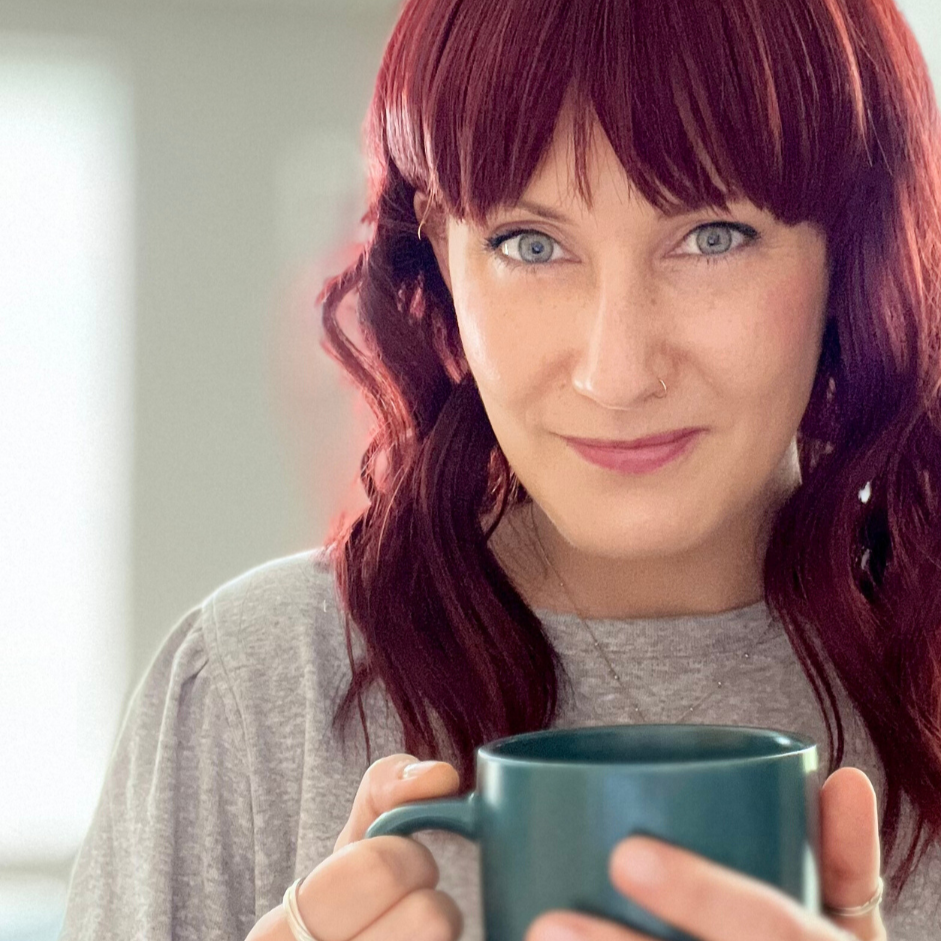 Jules, a woman with red hair and green eyes is holding a teal mug, smiling gently, in a well-lit room.