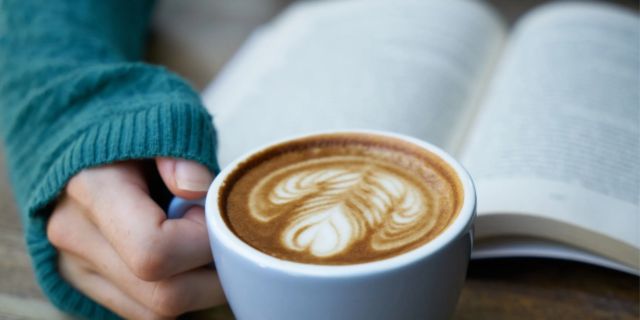 Person holding a white mug of latte with latte art, sitting next to an open book.